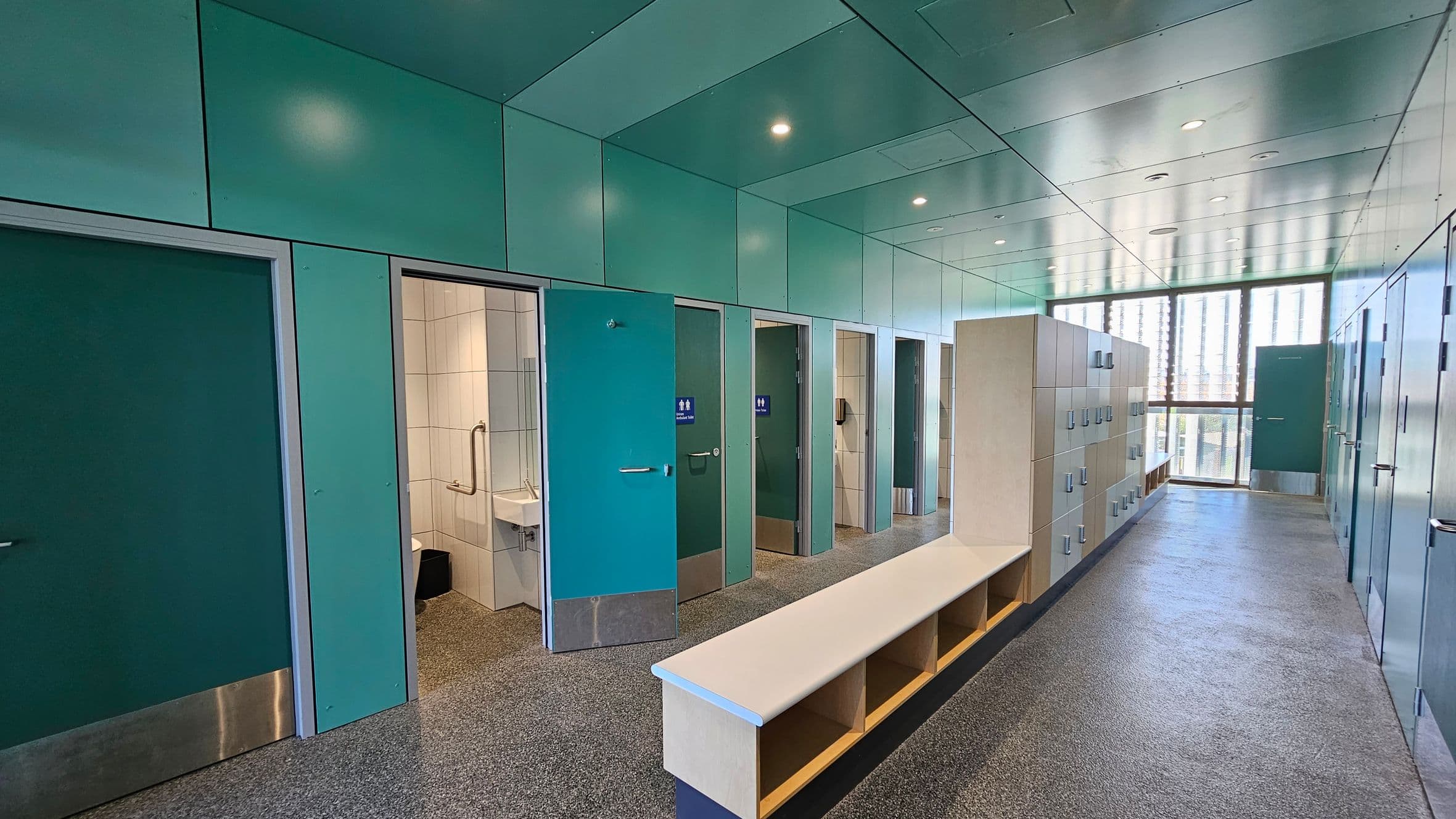 A modern public restroom with teal-coloured walls and ceiling. On the left, several stalls with teal doors, one open revealing a toilet and sink. In the centre, a long bench with open cubbyholes beneath. On the right, a row of lockers with silver handles. The floor is speckled grey, and the ceiling has recessed lighting. At the far end, large windows with vertical blinds allow natural light to enter.