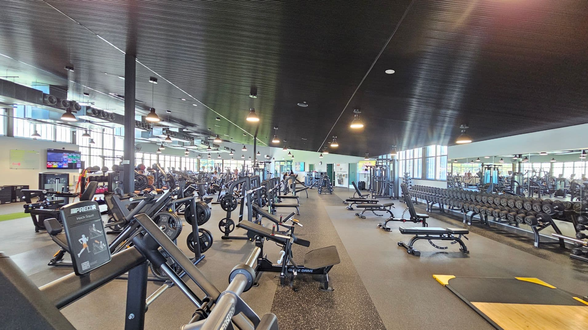 A spacious gym with a variety of exercise equipment. On the left, there are weight machines and a television mounted on the wall. In the centre, rows of benches and weightlifting equipment are neatly arranged. On the right, a long rack of dumbbells is positioned against a mirrored wall. The ceiling is dark with hanging lights, and the floor is covered with rubber mats. Large windows line the background, allowing natural light to fill the space.