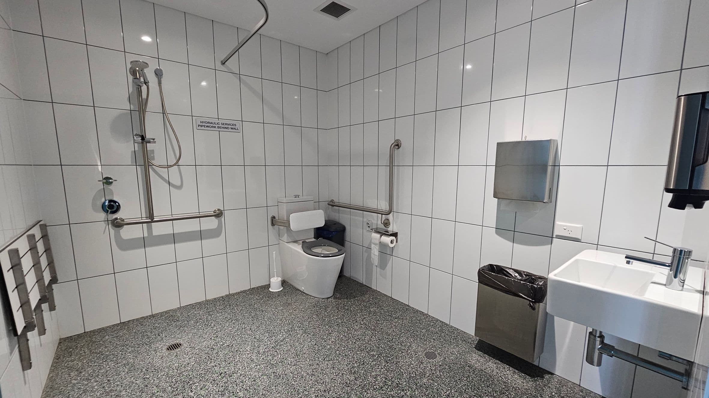 A spacious, accessible bathroom with white tiled walls and a speckled grey floor. On the left, there is a shower area with a handheld showerhead and a horizontal grab bar. In the centre, a toilet with support rails and a toilet roll holder is visible. On the right, a white sink with a modern tap is mounted on the wall, accompanied by a soap dispenser and a paper towel dispenser. A small waste bin is positioned below the paper towel dispenser. The ceiling is plain with recessed lighting.