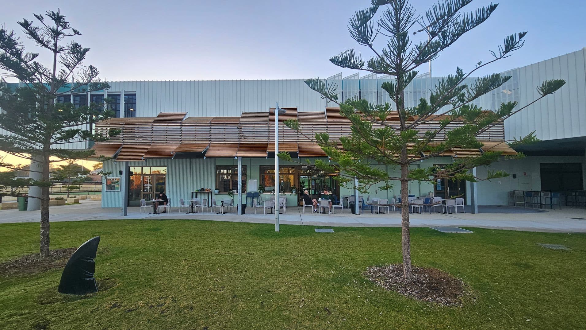 A modern building with a light grey facade and wooden slats on the upper section. In front, there is a patio area with tables and chairs, some occupied by people. The foreground features a grassy area with two young trees and a small sculpture. The building has large windows and a flat roof. The sky is clear, and the setting appears to be a public or commercial space.