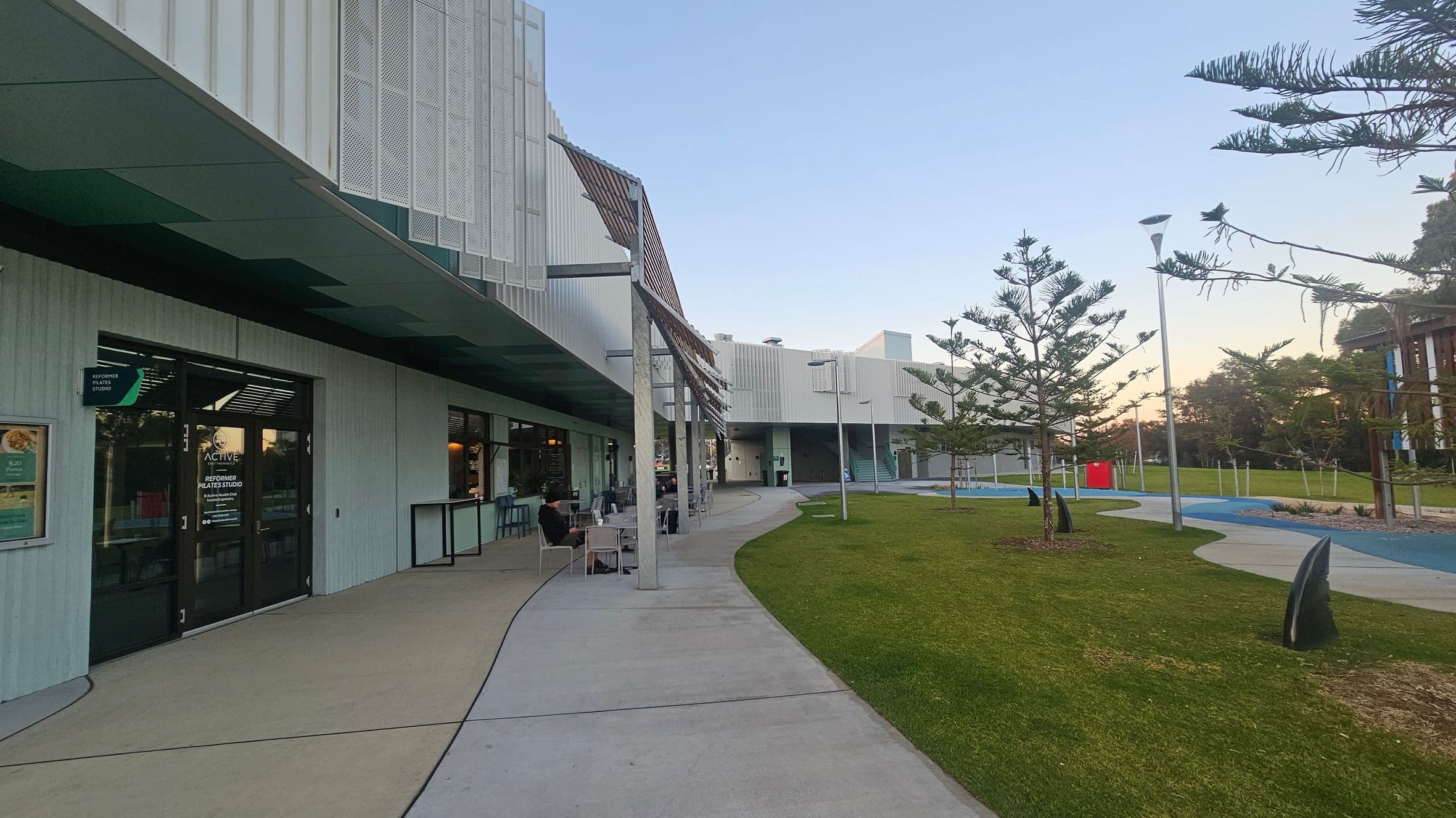 On the left, a modern building with a green overhang and large glass doors, featuring a sign for a Pilates studio. Several tables and chairs are set up outside, with a person seated. The right side has a landscaped area with grass, small trees, and a winding path. A few street lamps and a red bin are visible. The background shows more of the building with a staircase and additional greenery. The ground is a mix of concrete and grass.