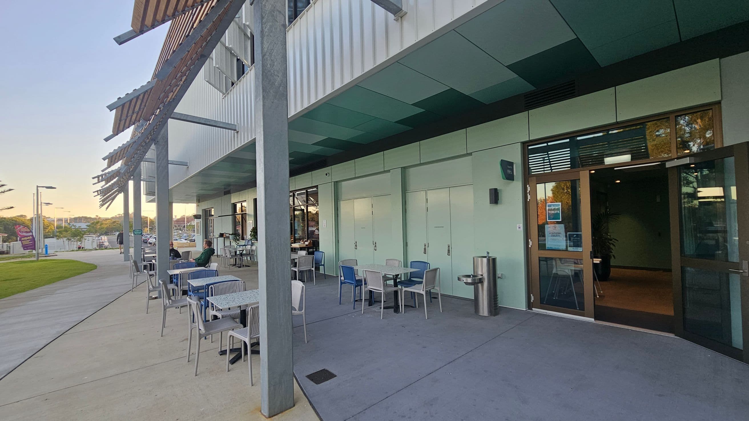 Outdoor seating area with several tables and chairs on a paved surface. On the right, a building with a glass door entrance and a water fountain nearby. The building has a modern design with a greenish hue. On the left, a grassy area and a pathway leading into the distance. Overhead, a series of angled metal beams provide partial shade. A few people are seated at the tables, and the area is well-lit with natural light.