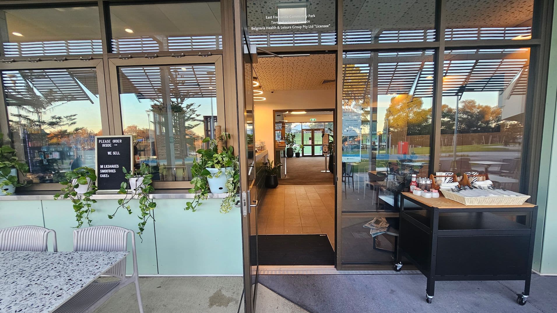 A café entrance with large glass doors and windows. On the left, a terrazzo table with light purple chairs is placed near a window ledge adorned with potted plants. A sign reads "Please order inside. We sell milkshakes, smoothies, cakes." On the right, a trolley holds condiments and utensils. Inside, a well-lit interior with a tiled floor and potted plants is visible. The ceiling has recessed lighting, and the background shows a grassy area through more glass doors.