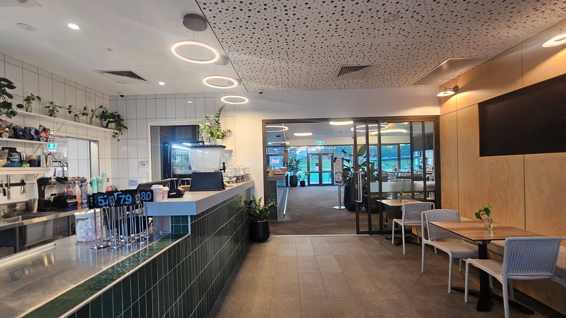 A modern café interior with a counter on the left featuring a display of order numbers and various items. Shelves above hold plants and other objects. The ceiling has circular lights and a perforated design. On the right, there are wooden walls with a black screen and a seating area with tables and chairs. The floor is tiled, and the background shows a glass door leading to an outdoor area with more plants visible.