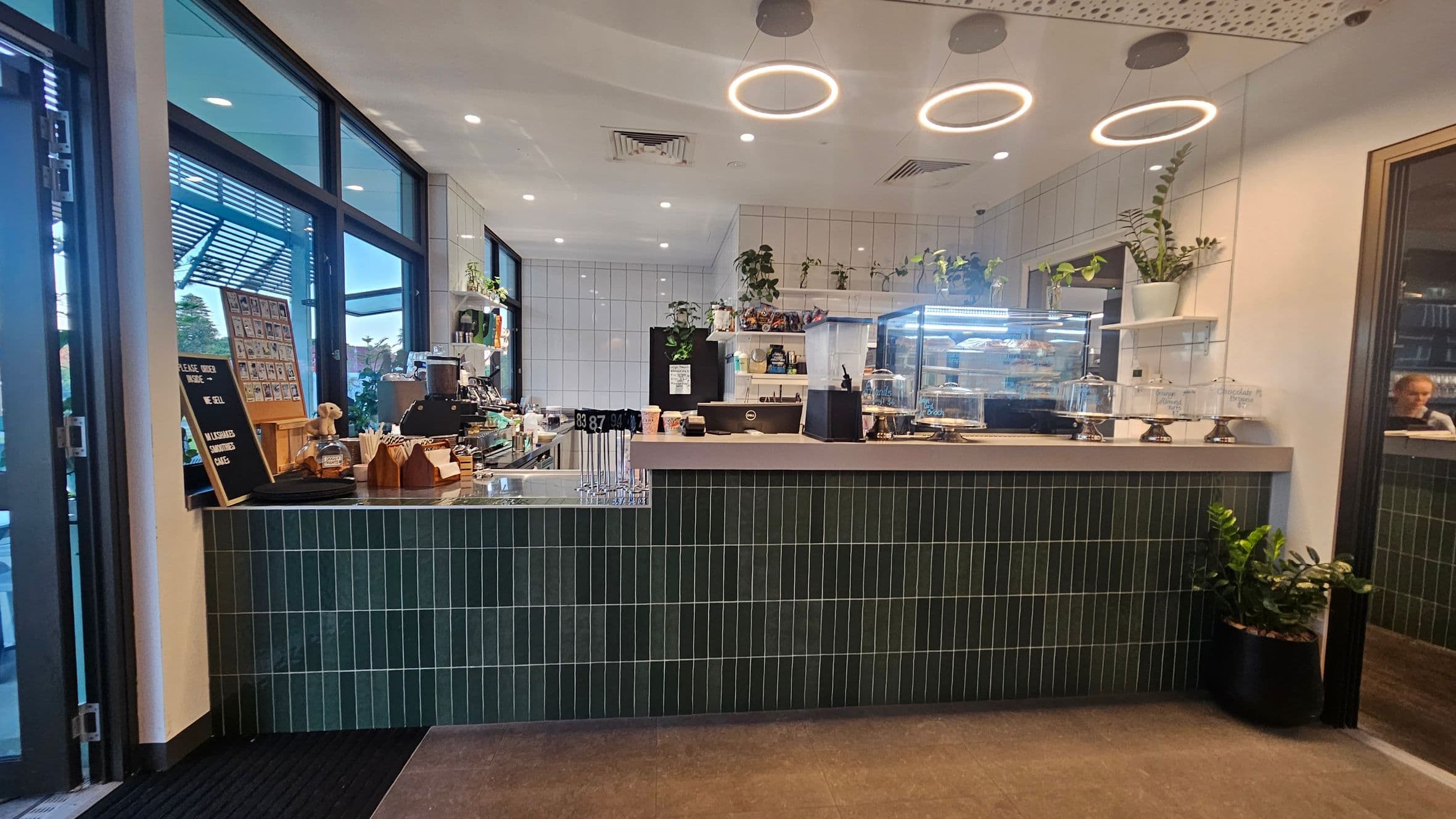 A modern café interior with a counter featuring dark green vertical tiles. On the left, large windows allow natural light in, with a menu board and various items on the counter, including a coffee machine and cups. The right side has a glass display case with pastries and cakes, and potted plants on shelves above. Circular ceiling lights illuminate the space, and a small plant is placed on the floor near the counter.