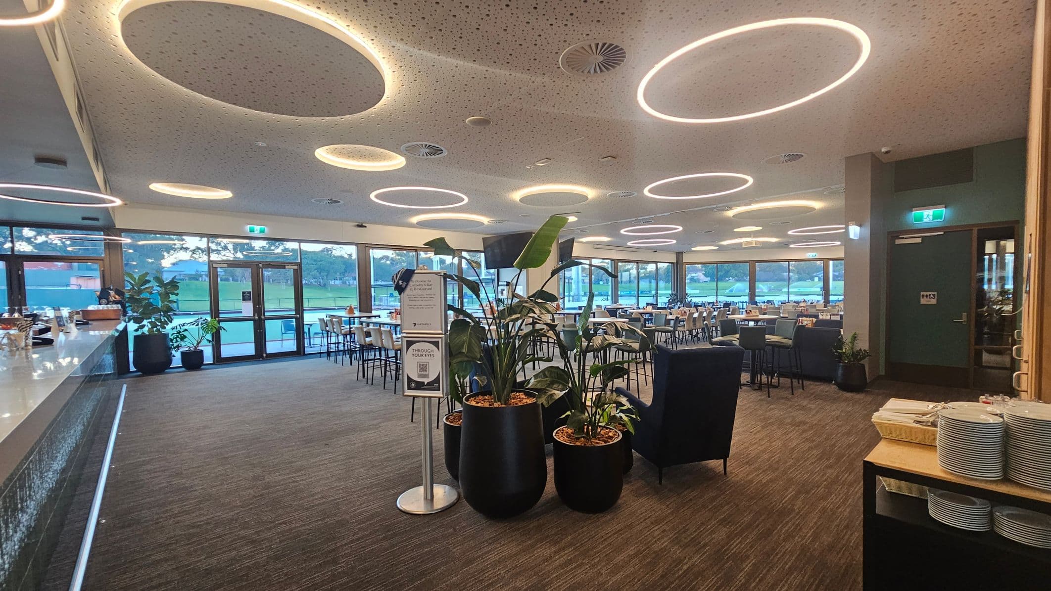 A modern indoor dining area with circular light fixtures on the ceiling. On the left, there's a long bar counter with stools. In the centre, large potted plants and a sign stand near a seating area with tables and chairs. The right side has a door with an exit sign above it and a shelf with stacked plates. Large windows in the background offer a view of a grassy outdoor area. The floor is carpeted, and the ceiling is white with circular patterns.