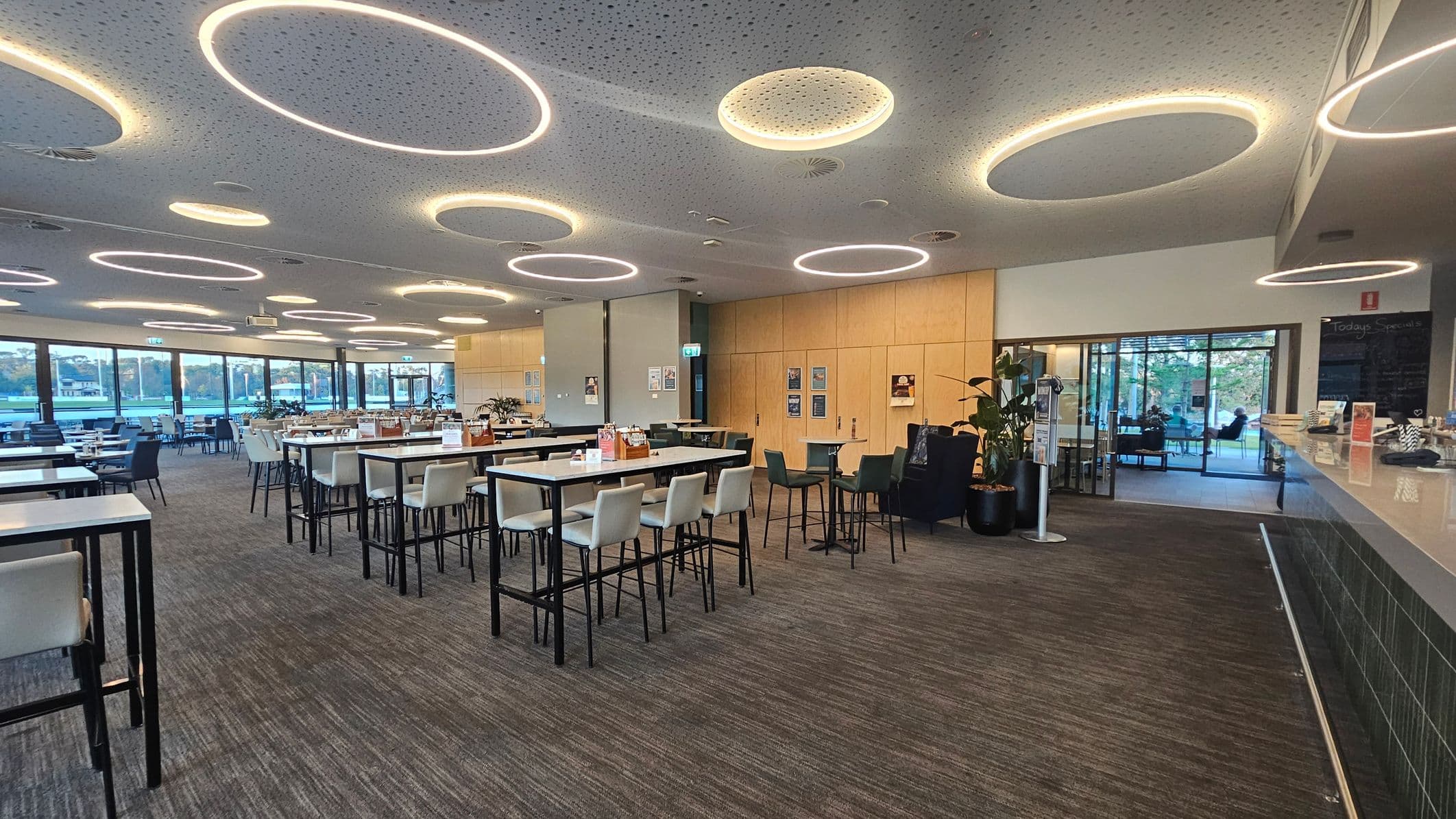 A spacious, modern dining area with high tables and chairs arranged in rows. The ceiling features circular light fixtures, creating a bright atmosphere. On the right, a counter with various items and a menu board is visible. The left side has large windows offering a view of the outdoors. The floor is carpeted in a grey tone, and the walls are light-coloured with some posters. In the background, there is a glass door leading to another seating area.