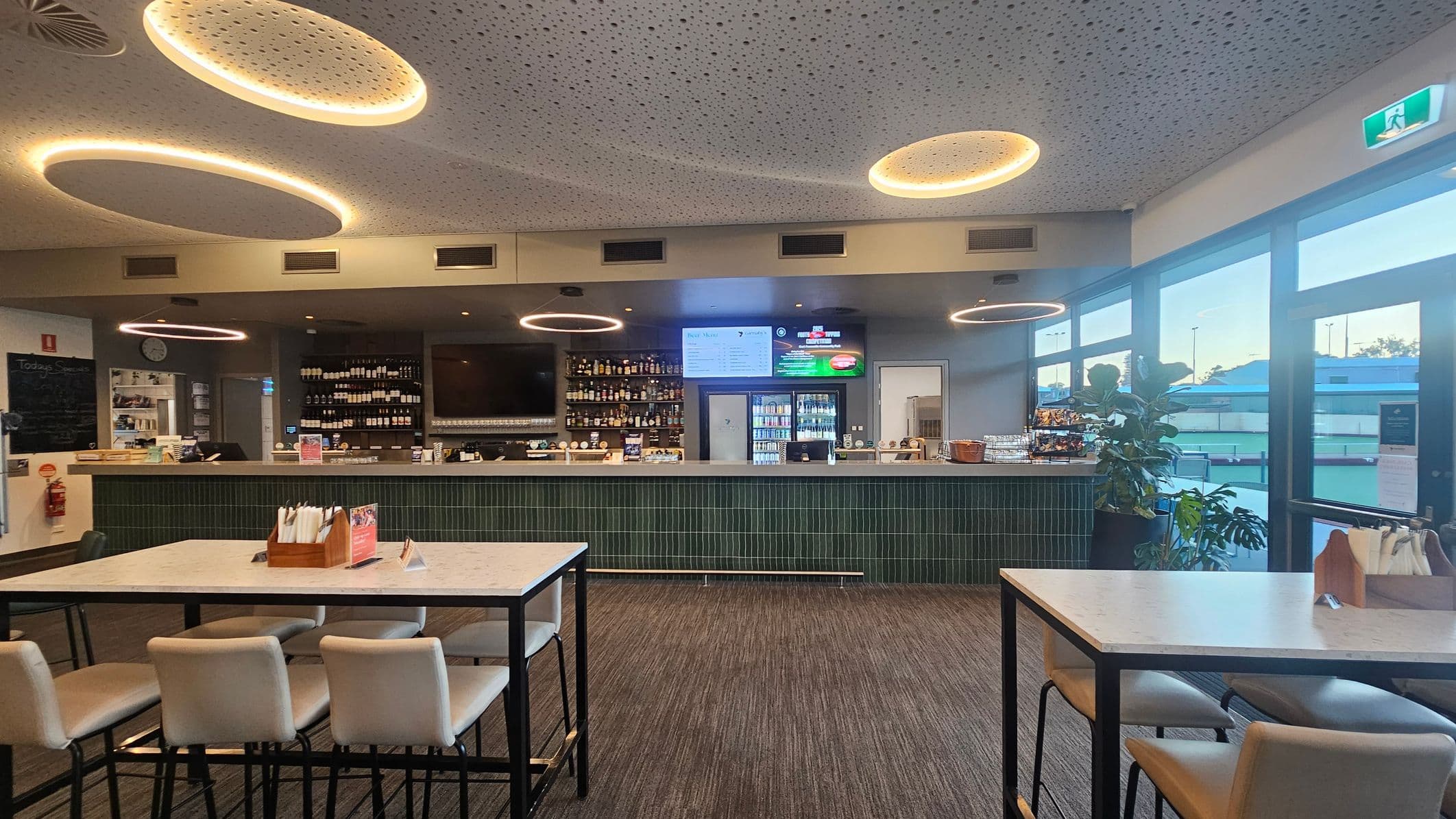 Interior of a modern bar with a long counter in the background, lined with bottles and a digital menu display. Circular light fixtures hang from a perforated ceiling. In the foreground, there are tables with white tops and cushioned chairs. To the right, large windows allow natural light in, with a view of the outside. A plant is placed near the window. The floor is carpeted in a neutral tone.