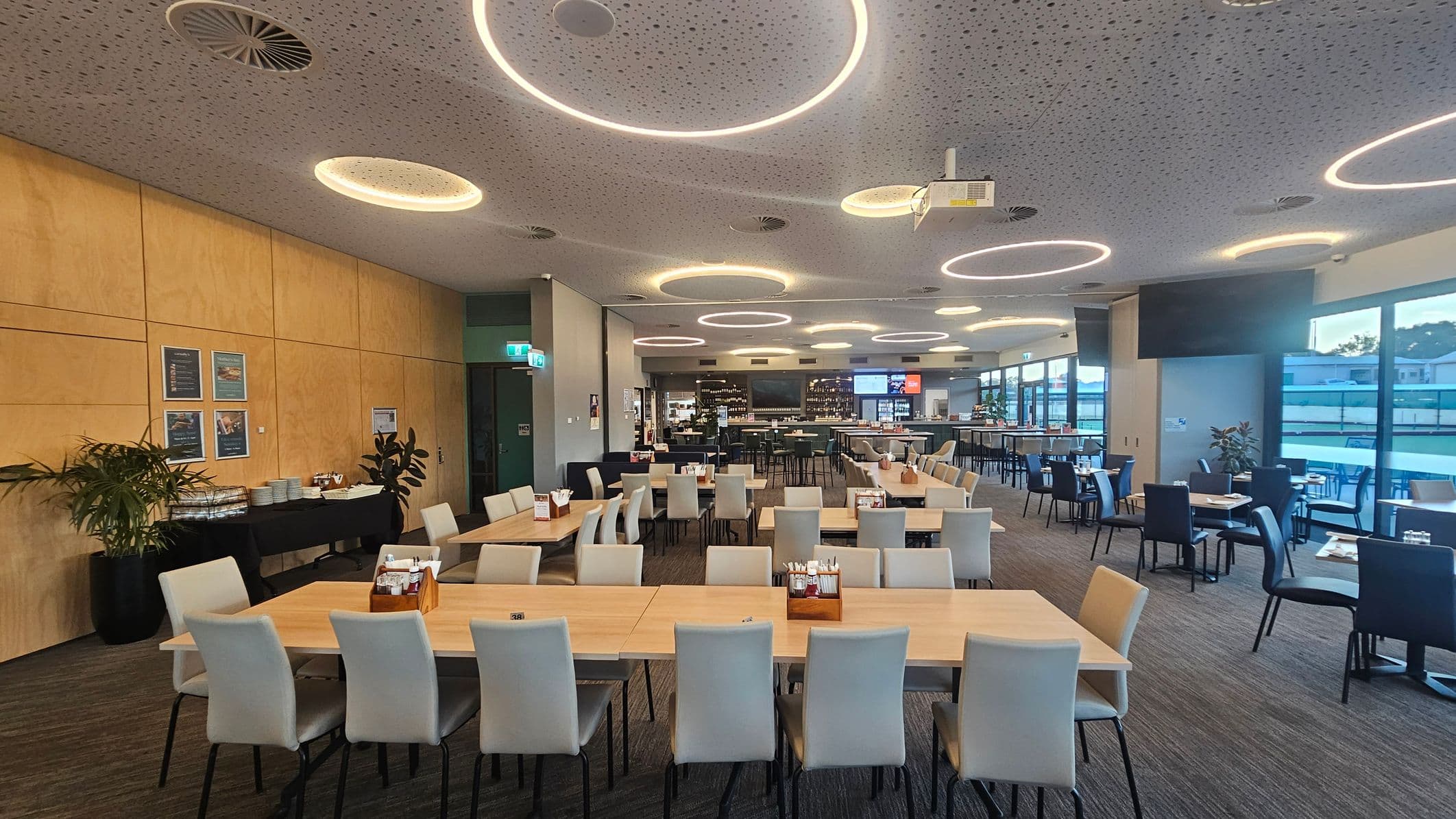 A spacious dining area with light wooden tables and white chairs arranged in rows. On the left, a wooden wall features framed pictures above a black table with plates and plants. The right side has large windows allowing natural light in, with more tables and chairs. The ceiling is dotted with circular lights and air vents. The floor is carpeted in a neutral tone. In the background, a bar area with shelves and digital screens is visible.