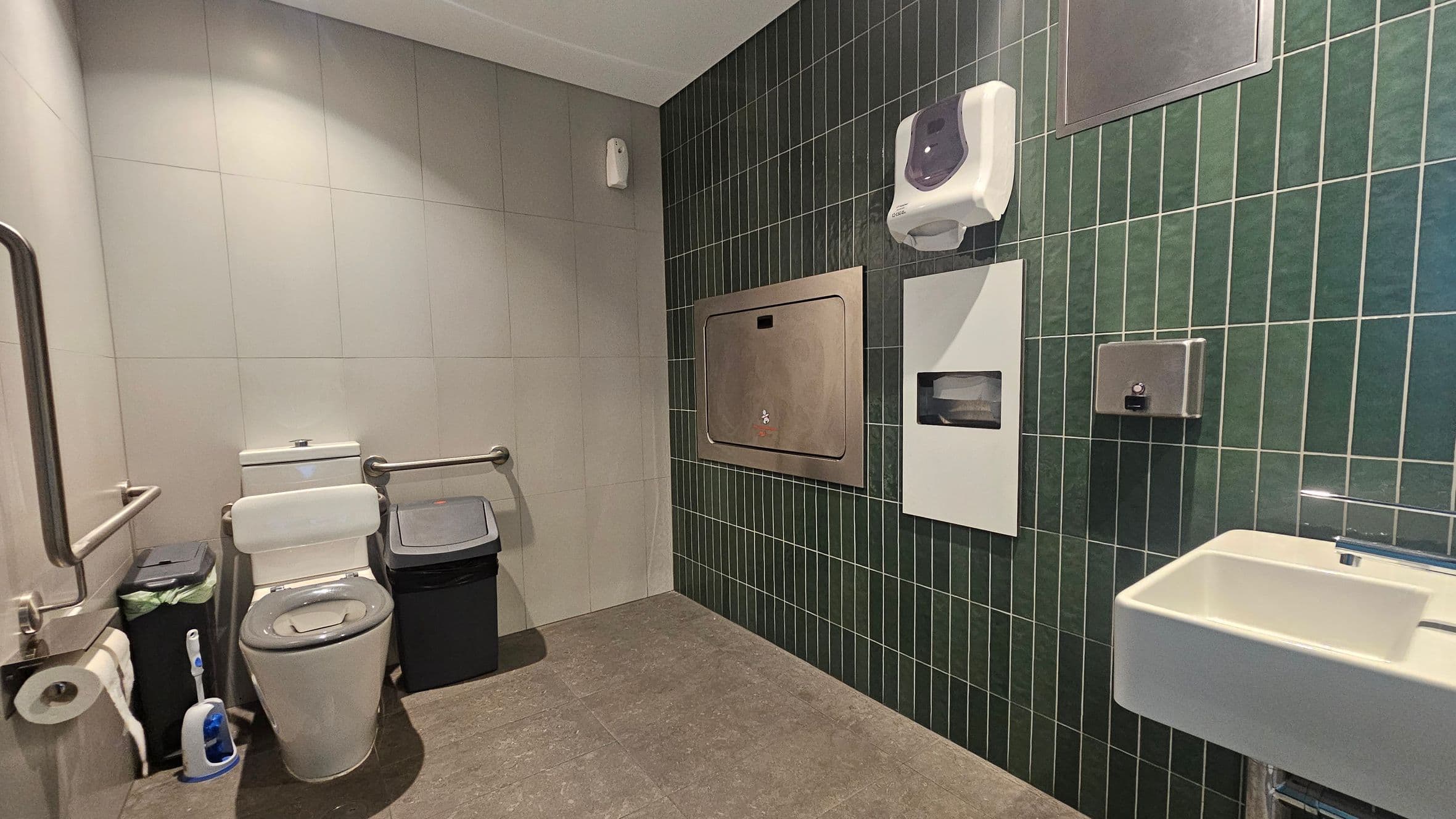 A public restroom with a toilet on the left, equipped with grab bars and a toilet brush nearby. A bin is placed next to the toilet. The right wall features dark green tiles and has a baby changing station, a soap dispenser, and a paper towel dispenser. A small white sink is mounted on the right side. The floor is covered with grey tiles, and the ceiling is light-coloured.