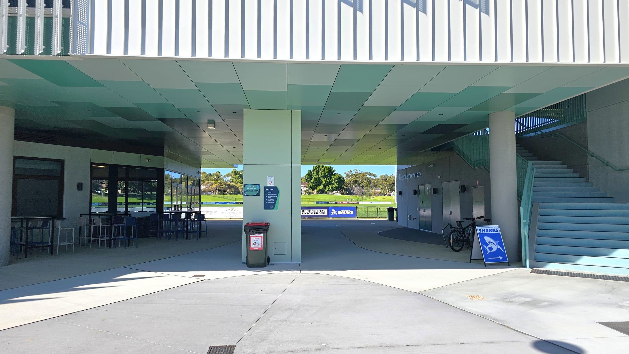 A modern building with a covered walkway. On the left, there are tables and chairs near large windows. In the centre, a green rubbish bin is placed against a pillar with signs. The background shows a grassy field with trees and advertising boards. On the right, a staircase leads upwards, with a bicycle parked nearby and a sign with a shark logo pointing right. The ceiling is flat and light-coloured, and the floor is concrete.