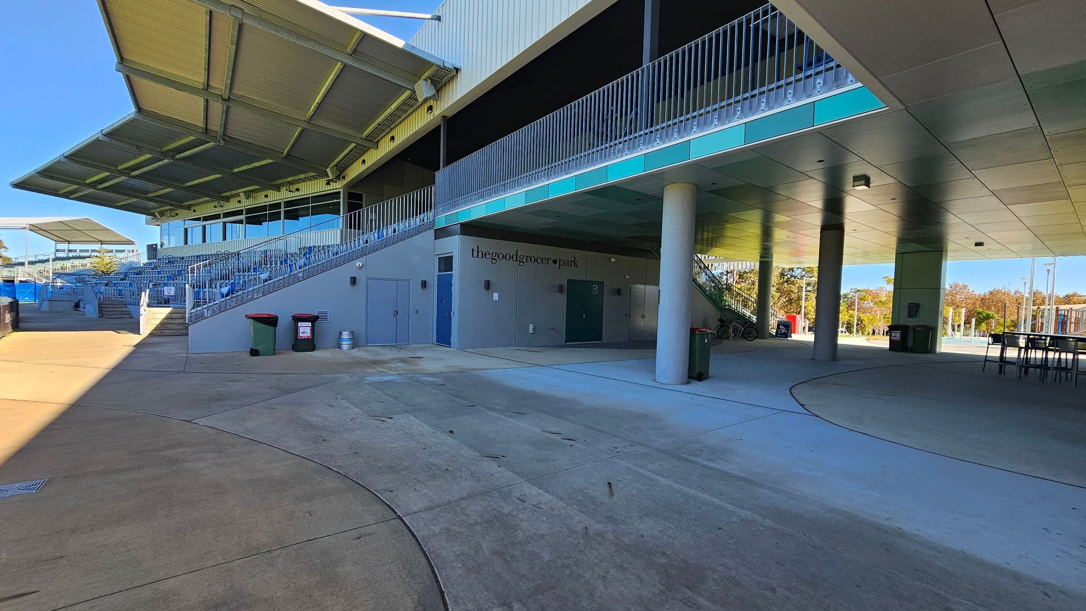 A sports stadium with a covered seating area on the left, featuring rows of blue seats and a large overhanging roof. Below the seating, there are several doors and a sign reading "thegoodgrocer park." The ground is a mix of concrete and paved surfaces. On the right, there are tables and bins, with a view of trees and open space in the background. The ceiling is flat with lighting fixtures, supported by columns.