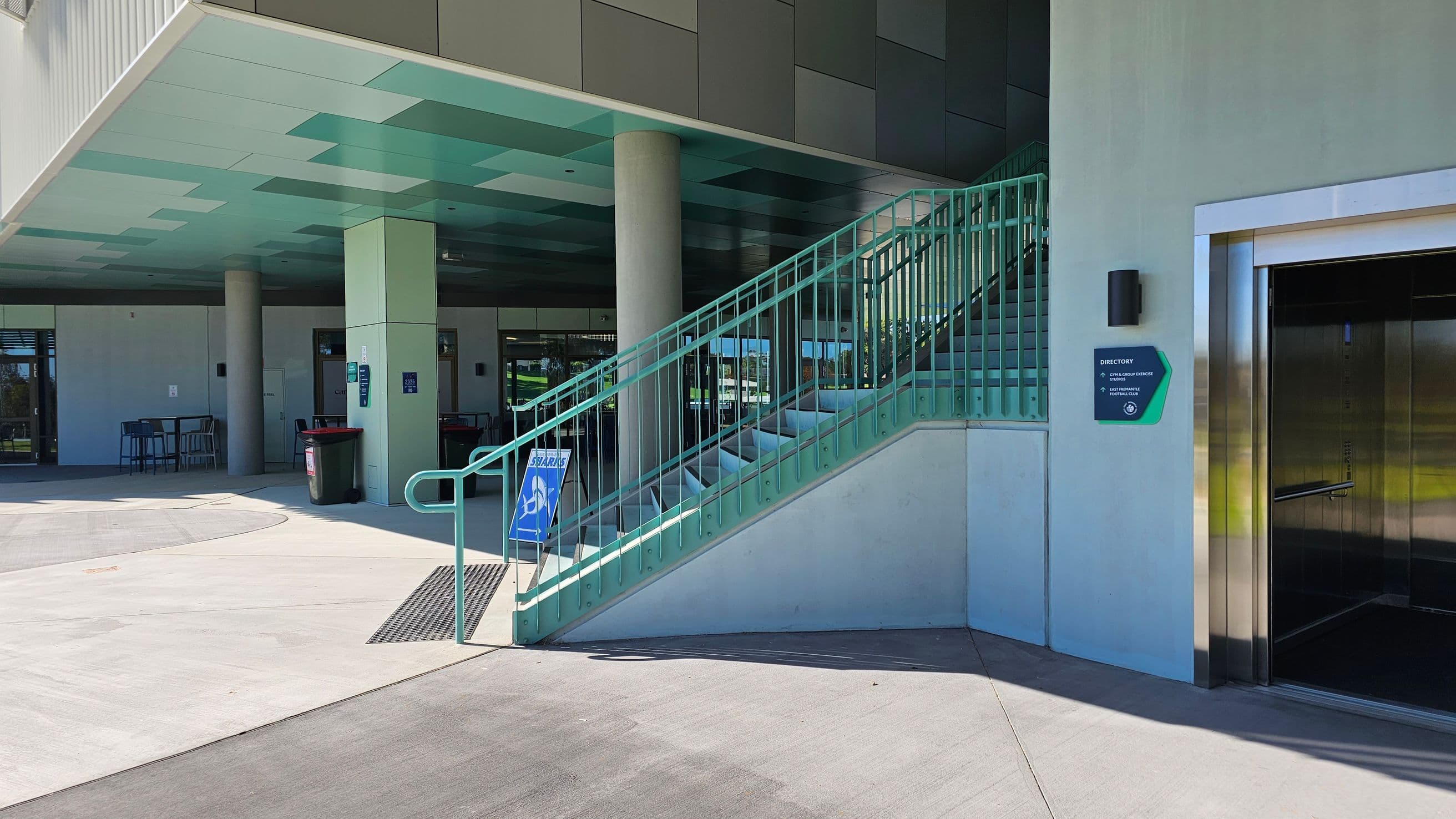A modern building entrance with a green and grey colour scheme. On the left, there is a staircase with a green railing and a sign indicating stairs. In the middle, a covered area with columns and a few tables and chairs. On the right, an open lift with a metallic finish and a directory sign on the wall. The floor is concrete, and the ceiling has a pattern of green and grey panels.