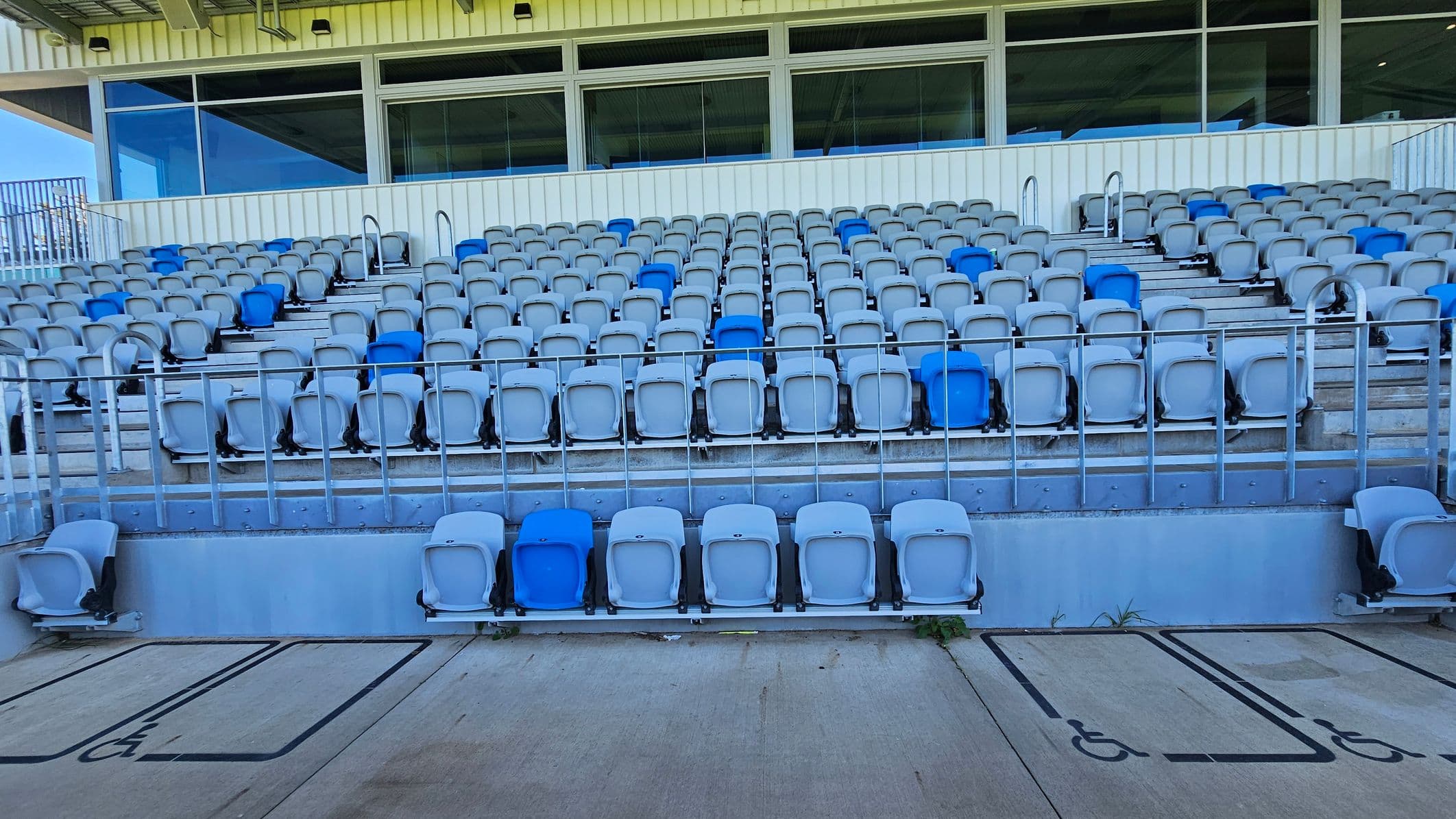 Rows of stadium seating with a mix of grey and blue plastic chairs. The seats are arranged in tiers, with metal railings separating sections. At the bottom, there are designated wheelchair spaces marked on the concrete floor. Behind the seats, there is a building with large windows and a white panelled exterior. The ceiling above the seating area is metal, with visible beams and lighting fixtures.