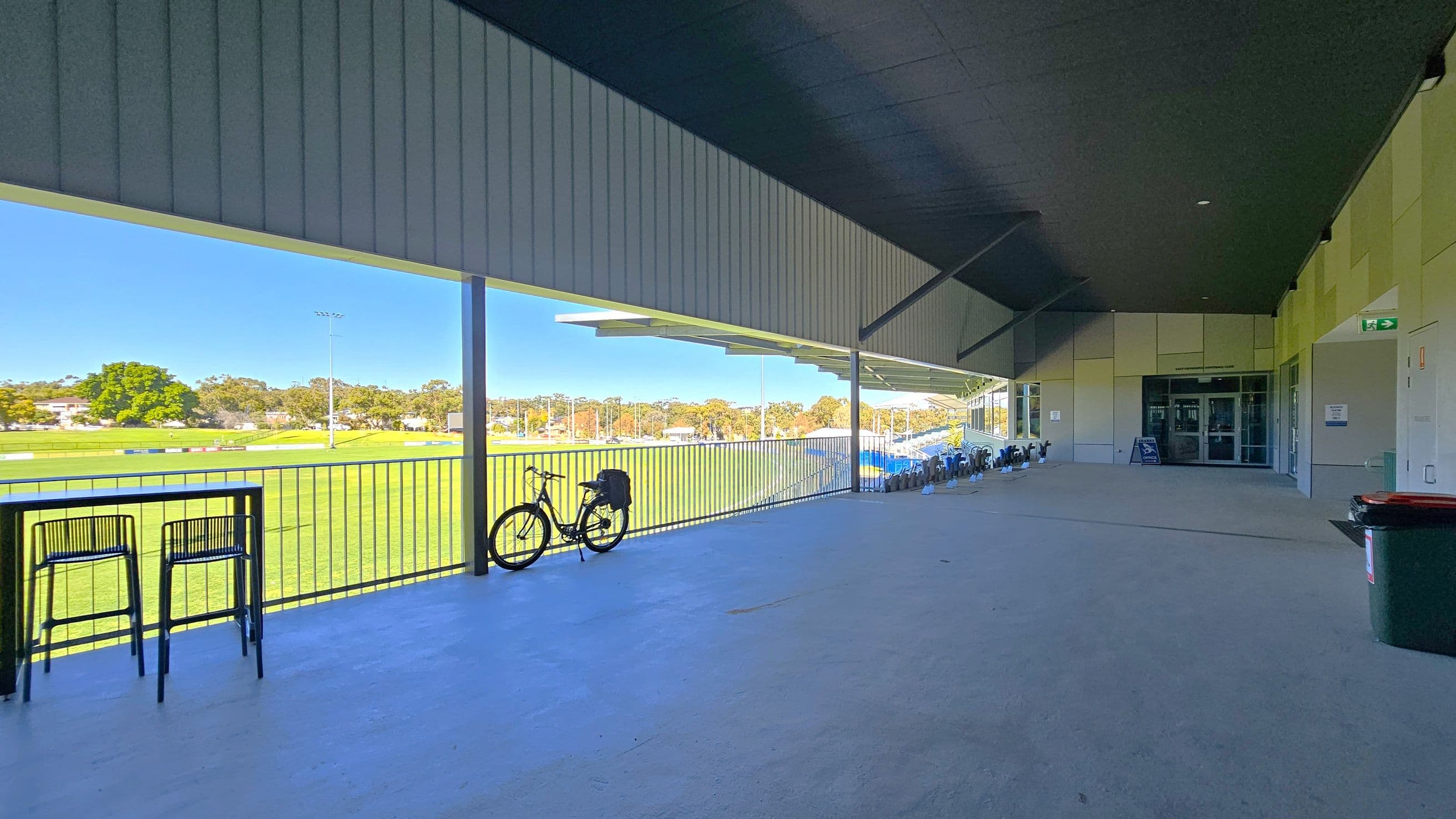 A covered outdoor area with a concrete floor and a dark ceiling. On the left, there is a railing overlooking a green sports field, with a bicycle leaning against it. Two high chairs and a table are positioned near the railing. On the right, a building entrance with glass doors and a sign is visible. A row of chairs is lined up along the wall, and a red-lidded bin is near the entrance. The background shows trees and a clear view of the field.