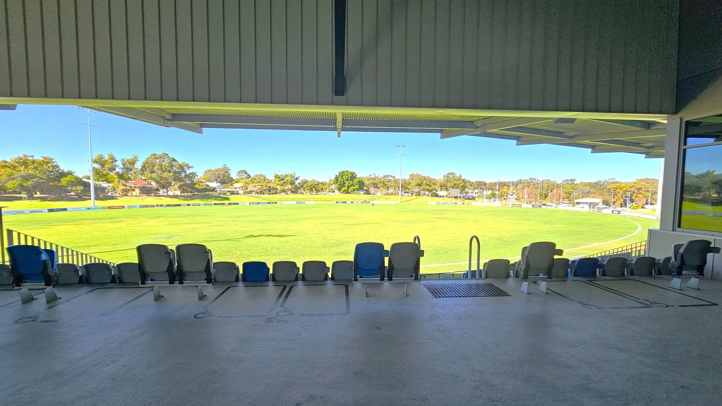 A view from a covered seating area overlooking a sports field. In the foreground, there are rows of empty seats with a concrete floor. The field is expansive and green, surrounded by a low fence. In the background, there are trees and some buildings visible beyond the field. The ceiling above the seating area is made of corrugated metal.
