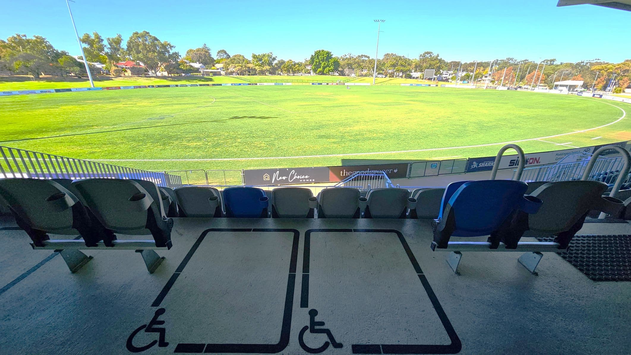 Rows of stadium seats in the foreground, with designated wheelchair spaces marked on the floor. Beyond the seats, a large, well-maintained grassy sports field is visible. Advertising boards line the perimeter of the field. In the background, there are trees and a few buildings, suggesting a park-like setting. The sky is clear, and the lighting indicates a sunny day.