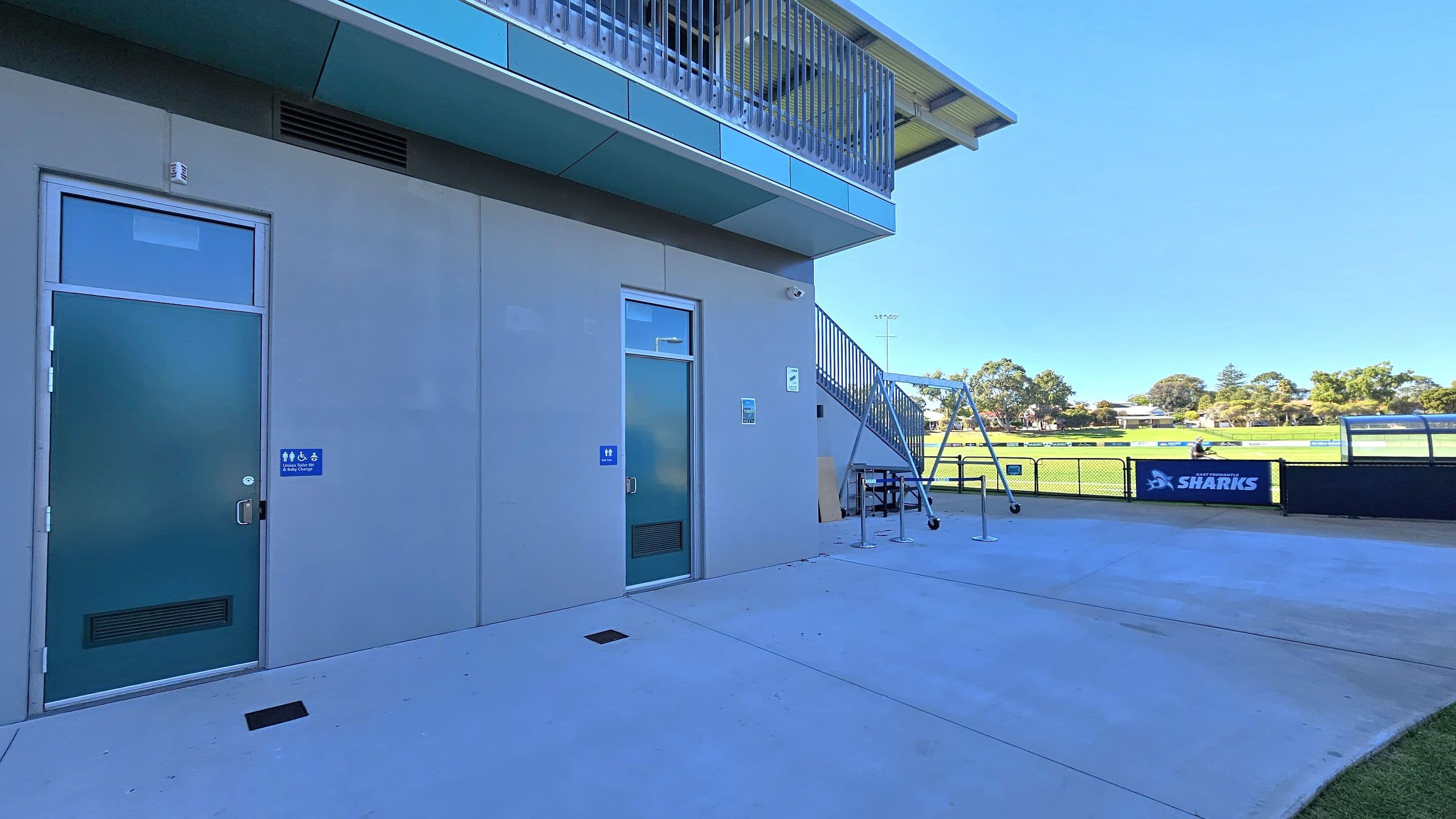 On the left, there are two doors with signs indicating accessible toilets. The building has a grey exterior with a blue overhang. In the middle, a staircase leads up to a balcony. On the right, a sports field is visible with a fence displaying a "Sharks" banner. The ground is paved with concrete, and there are some trees and houses in the background.