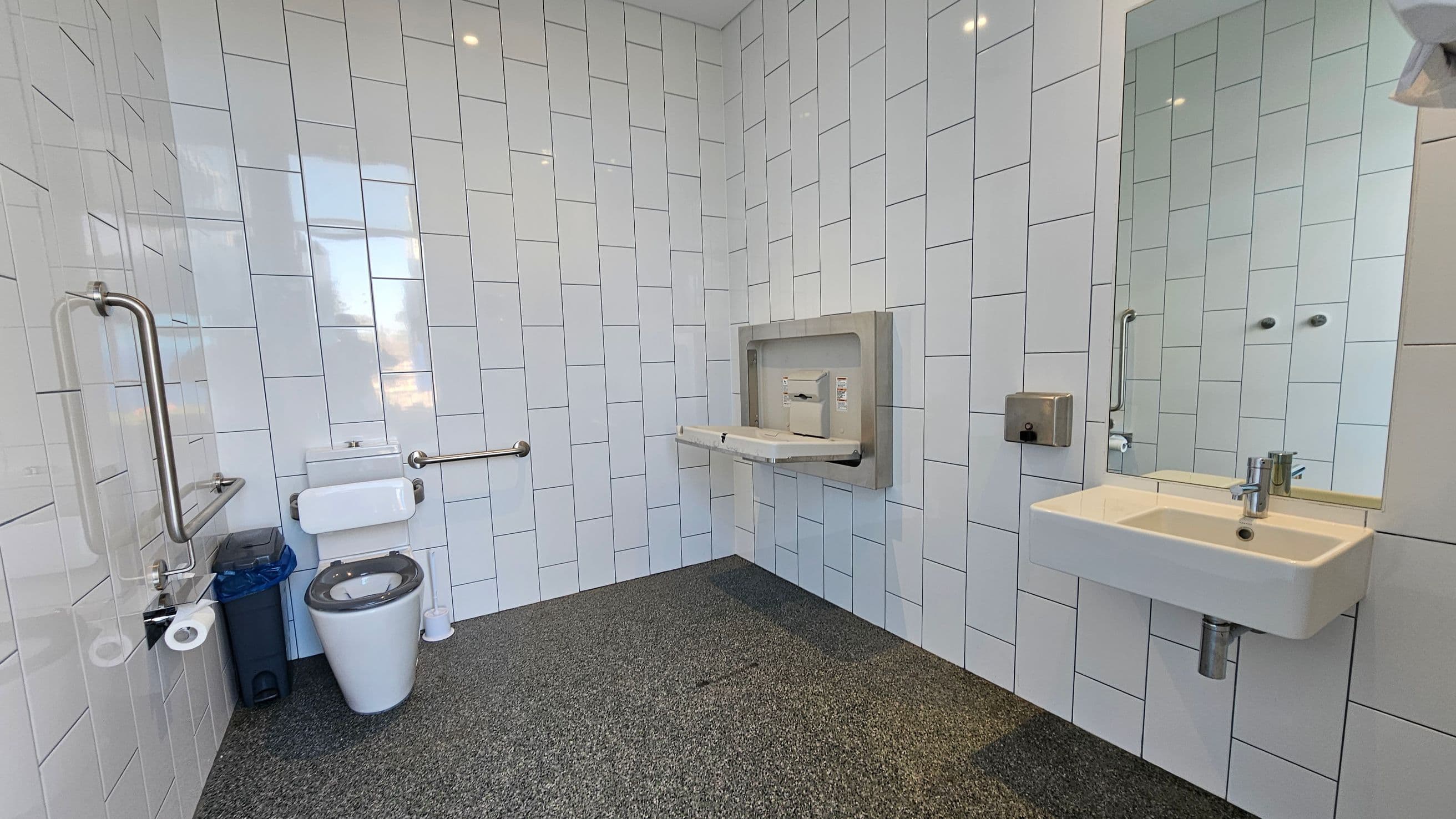 A public restroom with white tiled walls and a speckled grey floor. On the left, there is a toilet with a blue bin beside it and a grab rail. In the centre, a baby changing station is mounted on the wall. On the right, a white sink with a mirror above it and a soap dispenser to the side. The ceiling has recessed lighting.