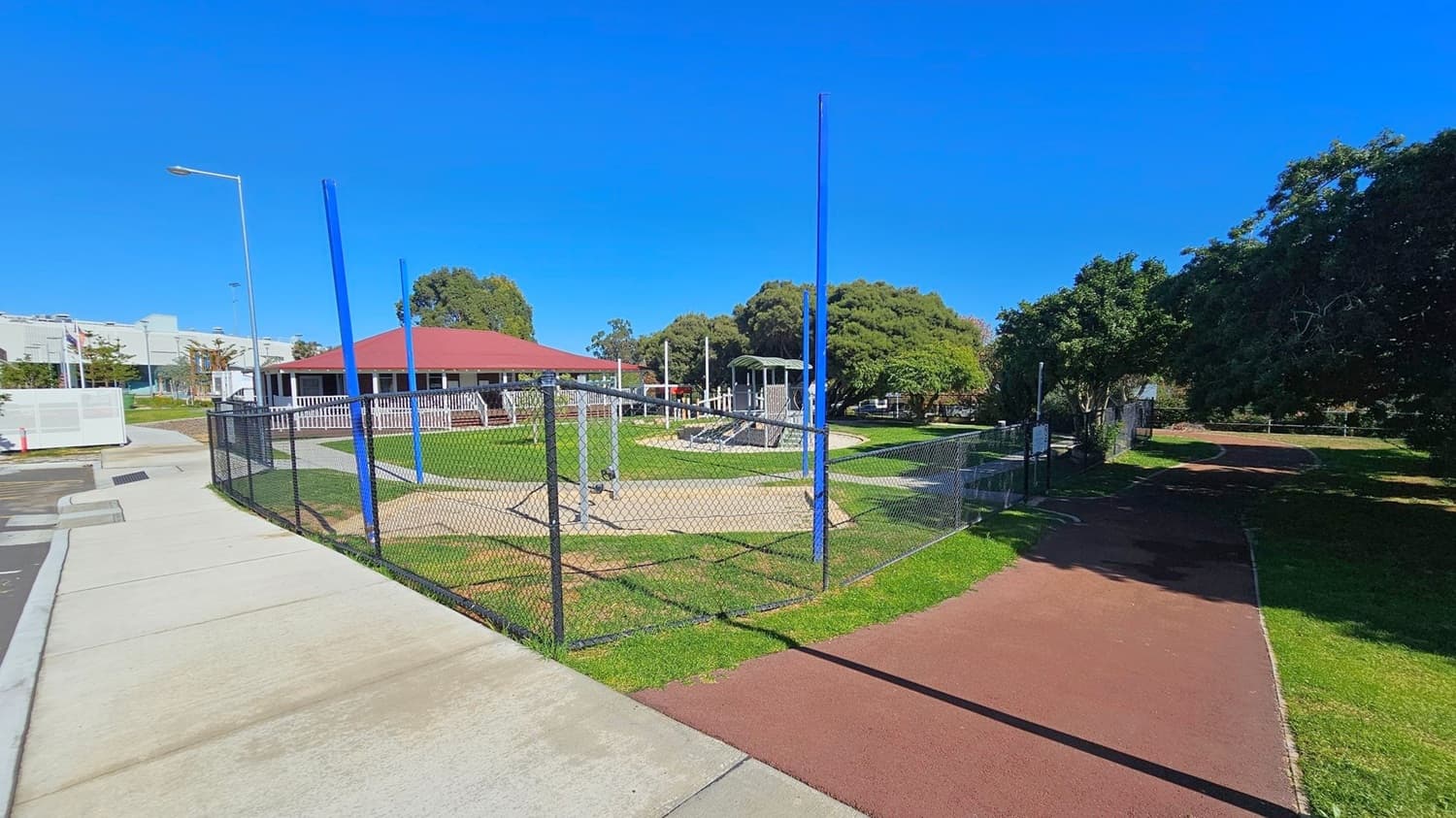 A park with a fenced playground area featuring blue poles and play equipment. To the left, there's a concrete path and a building with a red roof. In the background, trees provide shade, and a grassy area surrounds the playground. A red path runs alongside the fence on the right, leading into a more wooded area. A streetlamp is visible on the left near the path.