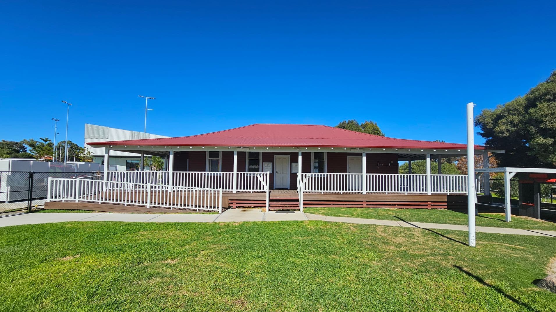 A single-storey building with a red roof and white railings is centred in the image. It has a wooden deck and a central door flanked by windows. The foreground features a grassy lawn with a concrete path leading to the entrance. On the left, there is a fenced area with some structures, and on the right, a large tree provides shade. The background includes a few tall poles and additional buildings.