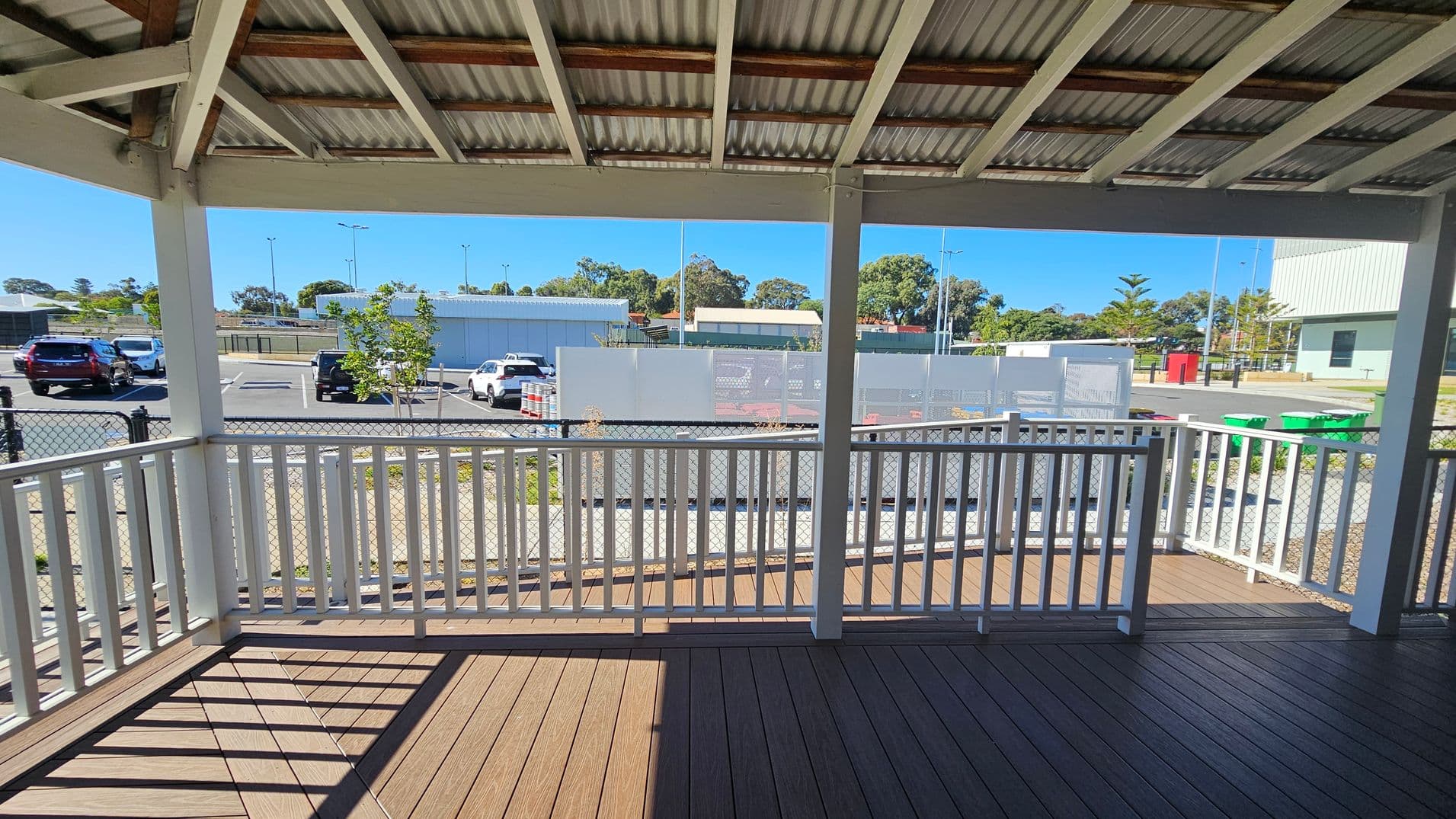 A covered wooden deck with white railings in the foreground. The ceiling is made of corrugated metal with wooden beams. Beyond the deck, a chain-link fence separates the area from a car park with several vehicles. In the background, there are buildings, trees, and a few streetlights. A few green bins are visible to the right.