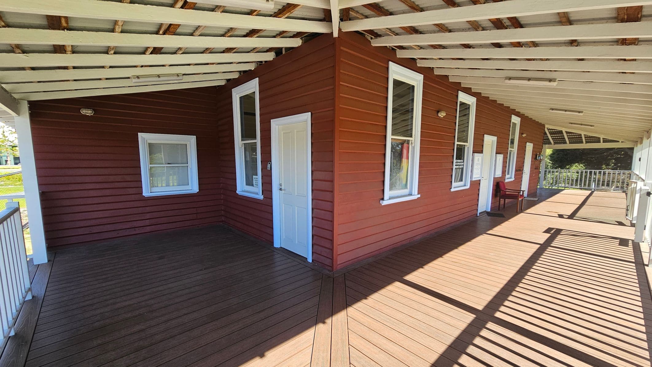 A wooden veranda with a white slatted roof and brown floorboards. The building has red siding with white-framed windows and doors. On the right, a chair is placed near one of the doors. The veranda is bordered by a railing, and sunlight casts shadows across the floor. The background shows greenery beyond the railing.