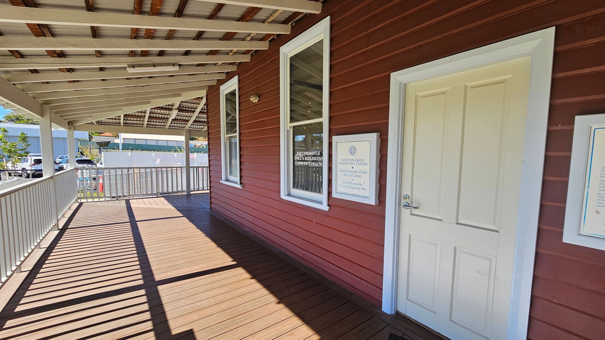 A wooden veranda with a brown floor and a white railing on the left. The building has red siding with two tall windows and a white door on the right. Signs next to the door read "East Fremantle Child & Adolescent Community Health" and "Stamford Green Community Centre." The ceiling is made of exposed beams and corrugated metal. In the background, there are parked cars and a modern building. Shadows from the railing create patterns on the floor.