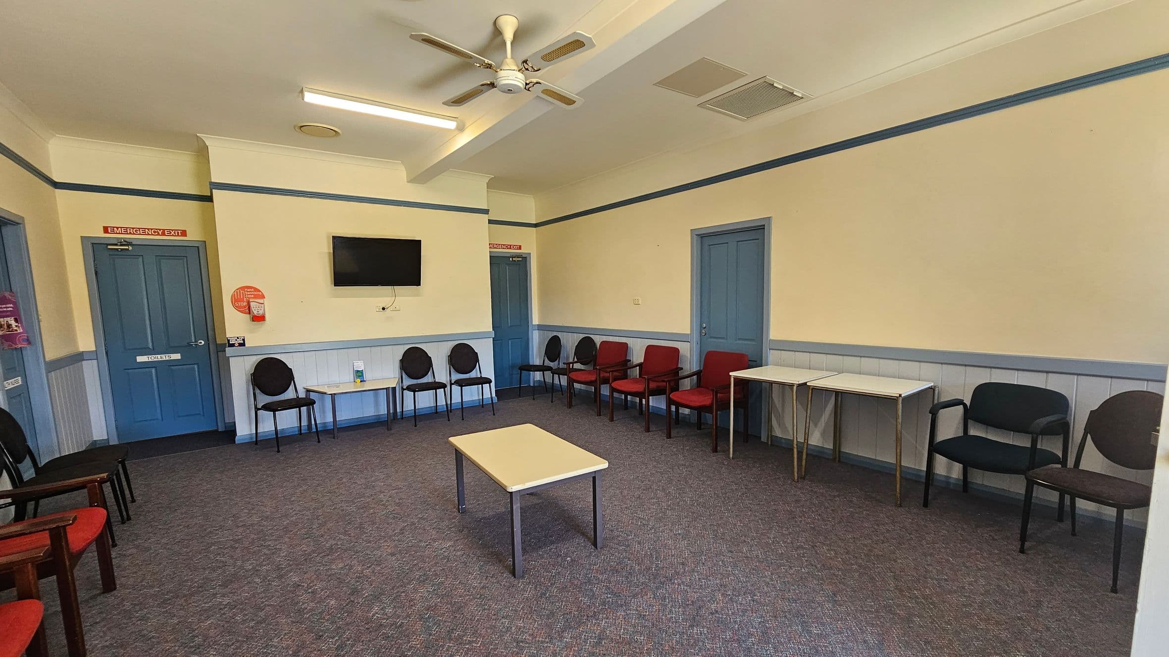 A waiting room with a purple carpet and cream walls. The ceiling is white with a ceiling fan and fluorescent lights. On the left, there are blue doors marked "Emergency Exit" and "Toilets," with black chairs and a small table beneath a wall-mounted TV. On the right, more black and red chairs are arranged along the wall, with two small tables. The room has a simple, functional design.