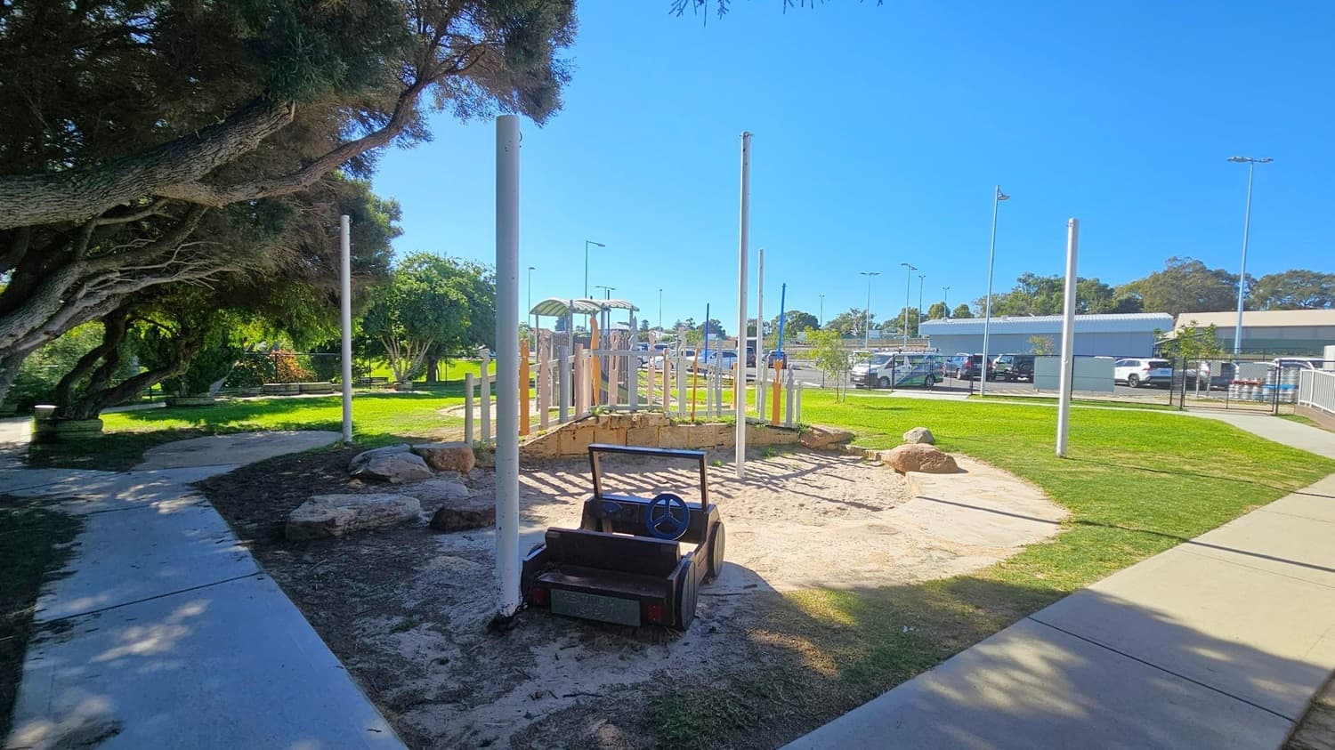 A playground area with a small, black toy car with a steering wheel in the foreground, set on sandy ground. To the left, there are large, leafy trees providing shade over a concrete path. In the middle, a wooden play structure is visible, surrounded by grass and rocks. The background features a parking area with several cars and a large building. The sky is clear and blue.