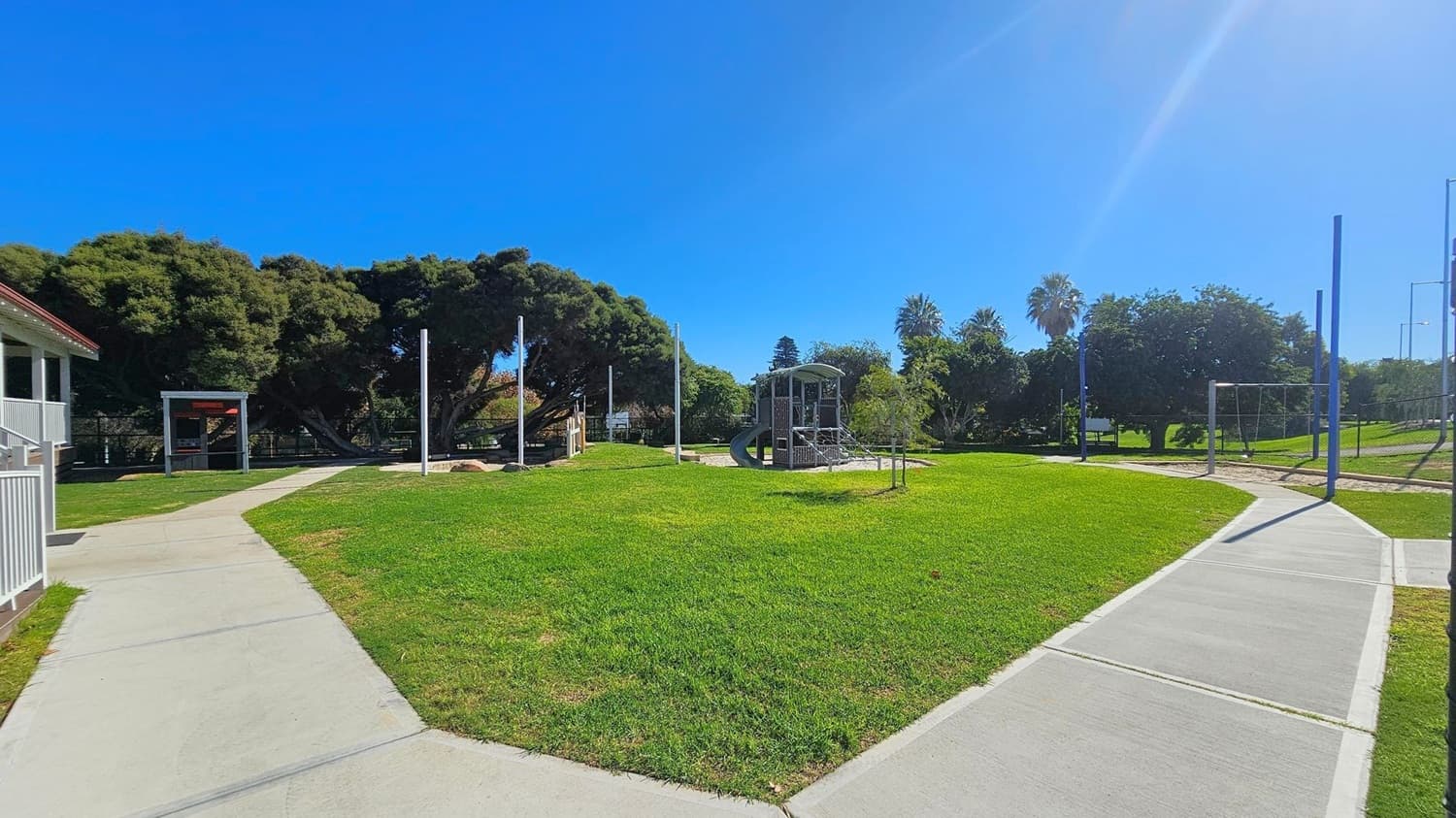 A park with a playground in the centre, featuring a climbing structure and slide. On the left, there's a building with a white railing and a small shelter. The right side has a swing set and a pathway leading further into the park. The background is filled with dense trees, including a prominent palm tree. The ground is covered with well-maintained grass, and the paths are paved with concrete.