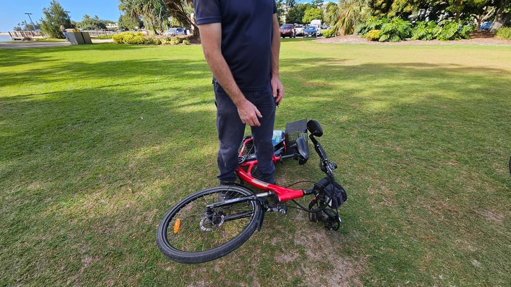 On the left, a man in a navy blue shirt and grey trousers is standing on grass. In the middle, a red electric bike is lying on its side with the handlebars and front wheel facing left. On the right, the bike's rear wheel and a black basket are visible. The background includes a grassy park area with trees, bushes, and parked cars.