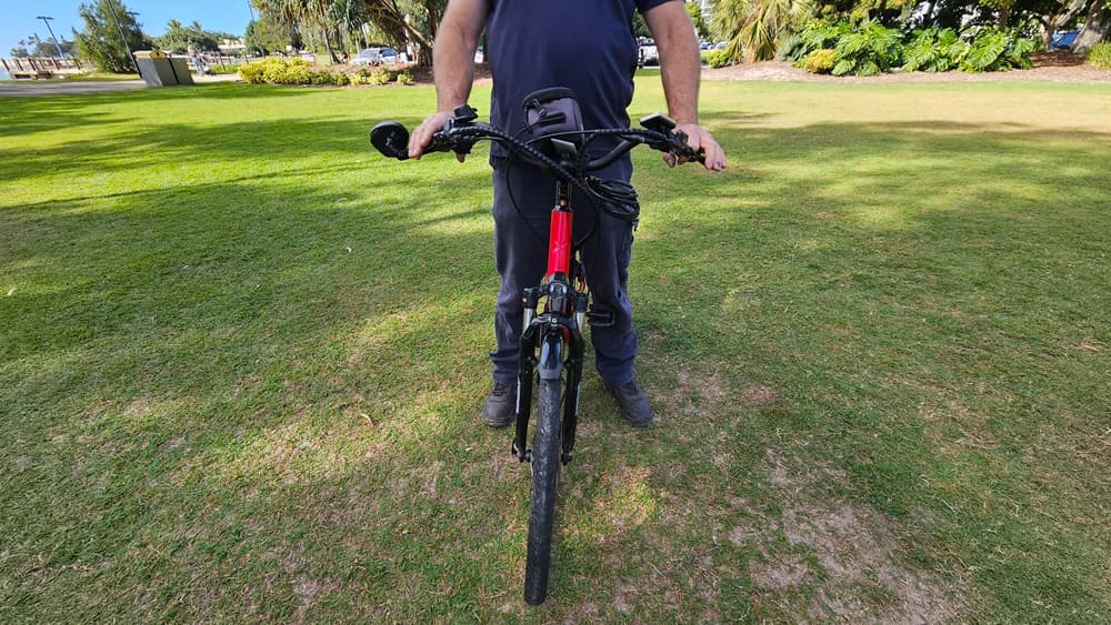On the left, a patch of green grass with some bushes in the distance. In the middle, a person wearing dark clothing is holding the handlebars of a red and black electric bike, standing on grass. The bike's front wheel is facing forward. On the right, more green grass and trees are visible. The background includes a grassy park area with trees and some distant structures.