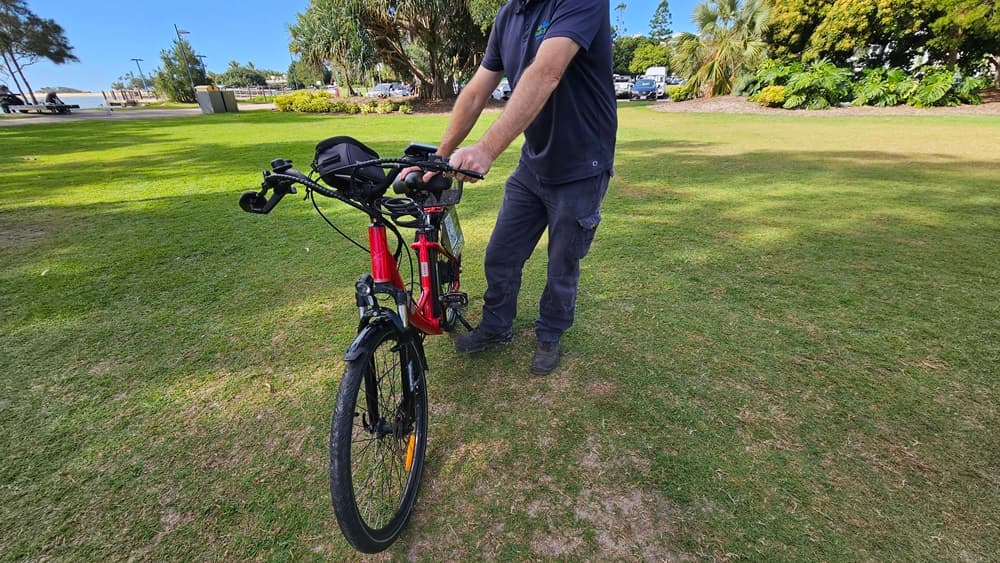 On the left, a patch of green grass with some bushes and trees in the distance. In the middle, a person wearing a dark shirt and trousers is holding the handlebars of a red and black electric bike, standing on grass. The bike's front wheel is facing forward. On the right, more green grass, trees, and parked cars are visible. The background includes a grassy park area with trees, bushes, and distant structures.