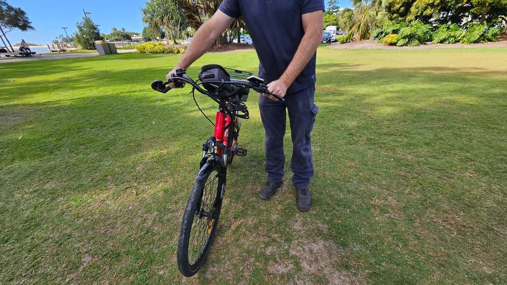 On the left, a grassy area with a few scattered trees and a distant building. In the middle, a person wearing a dark blue shirt and trousers is holding the handlebars of a red and black electric bike, standing on grass. The bike's front wheel is facing forward. On the right, more grass, trees, and a few parked cars are visible. The background includes a grassy park area with trees and bushes.