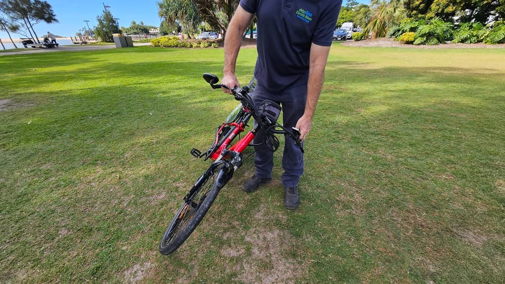 On the left, a grassy area with a few scattered trees and a distant pathway. In the middle, a person wearing a dark blue shirt and trousers is holding a red and black electric bike by the handlebars, lifting its front wheel off the ground. On the right, more grass, trees, and a few parked cars are visible. The background includes a grassy park area with trees and bushes.