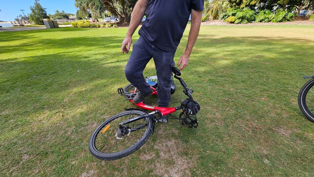On the left, a red electric bike lies on its side on a grassy area. In the middle, a person wearing a dark blue shirt and trousers is standing with one foot on the bike's frame and holding the handlebars. On the right, part of another bicycle wheel is visible on the grass. The background includes a grassy park area with trees, bushes, and a distant pathway.