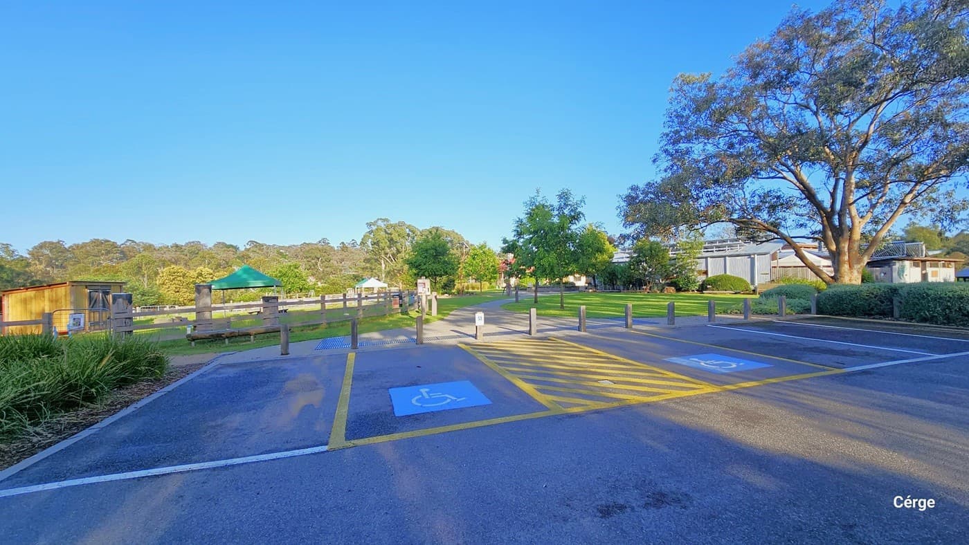 A concrete pathway leading to the venue, surrounded by well-maintained grass. Along the right side of the path stands a concrete structure resembling a horse's head. Further ahead, the venue's building with a corrugated design is visible, accompanied by other structures, street lights, and tall trees.