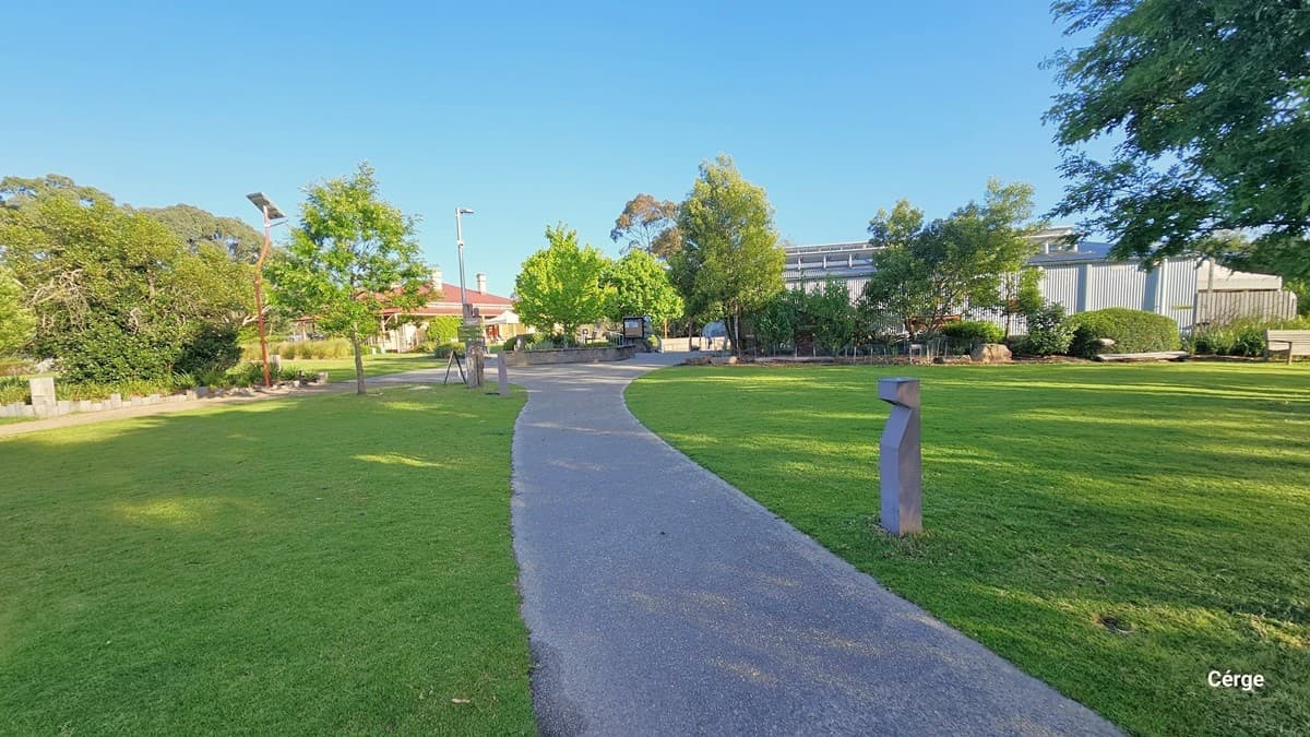 Two accessible car parking spaces with yellow markings and rectangular bollards are in place. A pedestrian driveway leads guests onto the premises. On the left stands a yellow horse stable, enclosed by a fence. Signage is visible, and to the right, a tall tree provides shade to part of the parking area. The surroundings are adorned with well-maintained shrubs, plants, and green grass.
