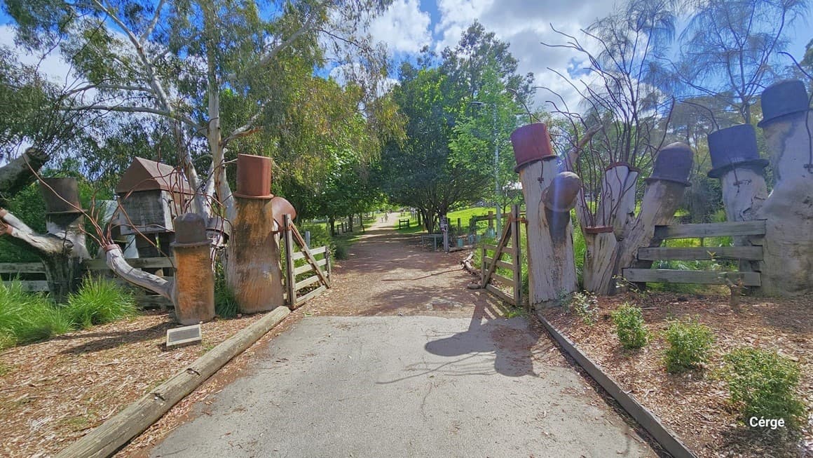 A concrete pathway leading to the farm through an open set of wooden gates. Both sides of the pathway are bordered with concrete and ornamental plants and bollards. On both sides, there are concrete sculptures with hats and a brick house. Tall trees surround the area.