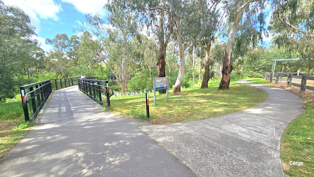 A lengthy bridge with paired black metal railings. The ground surface is composed of non-slip material, marked by a straight yellow line. Prior to the bridge entrance, a concrete pathway branches to the right over uneven terrain. A signage is positioned before the bridge entrance. The surroundings include towering mature trees, bushes, and tall grass.