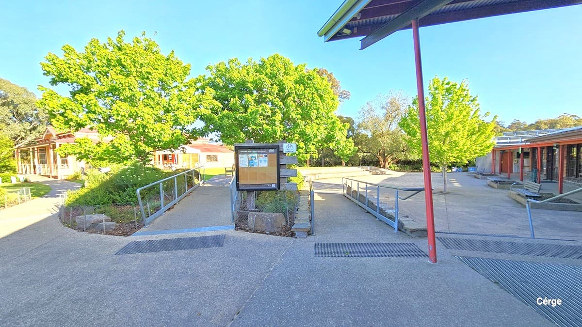 A courtyard with a central notice board surrounded by pathways and railings. On the left, there are buildings with red roofs partially obscured by lush green trees. On the right, more buildings with similar architecture are visible, along with additional trees. The ground is paved, and the area is well-lit, suggesting a sunny day. A metal roof structure is partially visible at the top right corner.