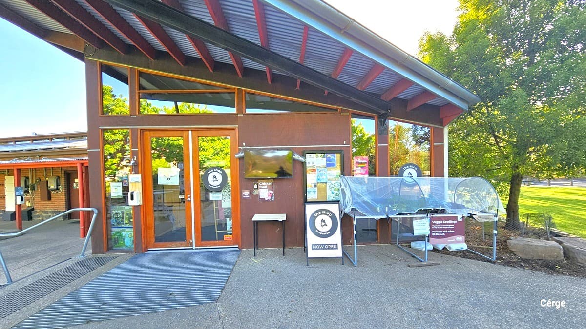 A building entrance with large glass doors and windows. On the left, there's a metal handrail leading up to the entrance. The doors have various signs and notices. To the right, a covered display or table with more signs and a small board indicating a shop is open. The building has a slanted metal roof with visible beams. In the background, there are trees and a grassy area, suggesting a park-like setting. The ground is paved with a textured surface.