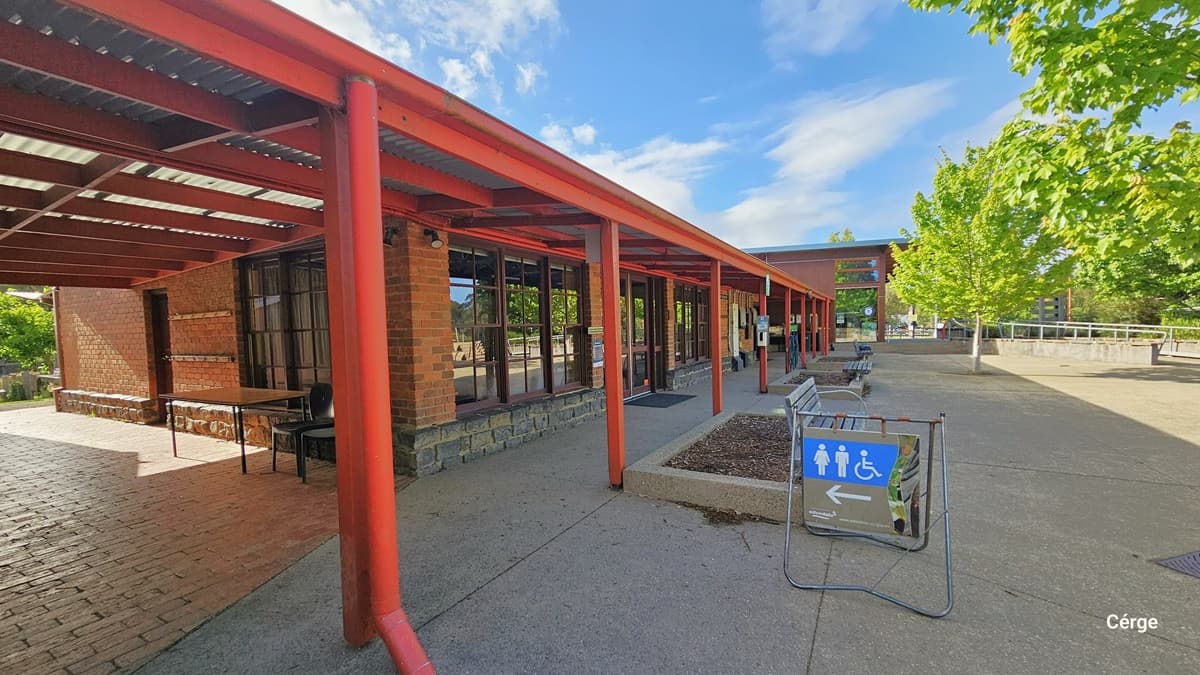 A walkway with a red metal pergola on the left, covering a brick building with large windows. A table and chair are placed near the entrance. On the right, there is a paved area with a sign indicating directions to accessible toilets. Several benches and a row of green trees line the path. The sky is partly cloudy, and the area is well-lit by natural light.