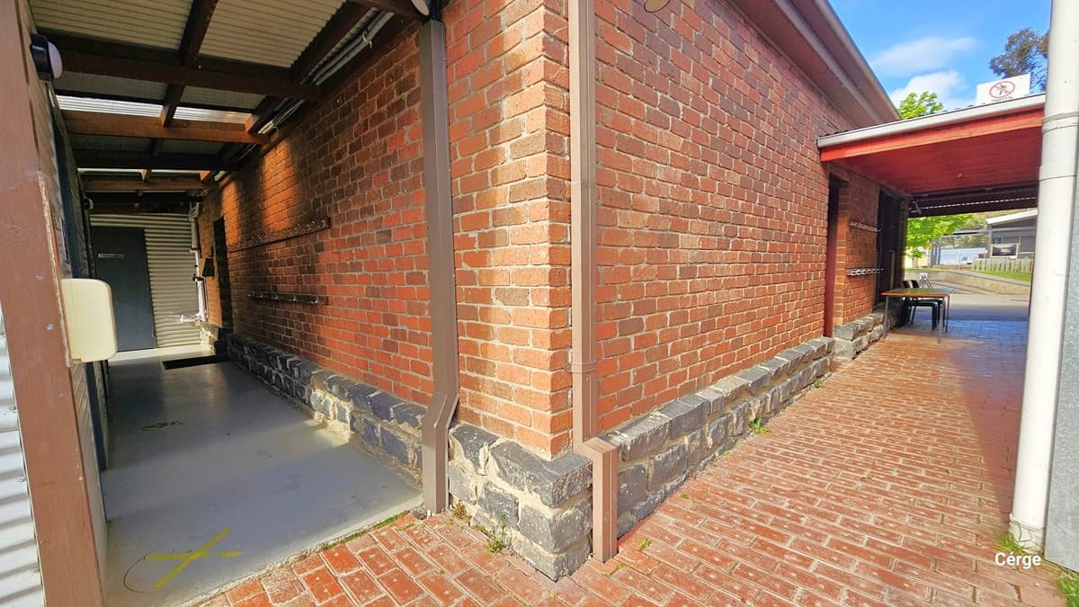 A narrow alleyway with a red brick wall on the right and a covered walkway on the left. The floor is made of red bricks, and the ceiling of the walkway is supported by metal beams. At the end of the alley, there is a table and chairs. The base of the brick wall is lined with dark stone. In the background, there are trees and a building visible through the open end of the alley.