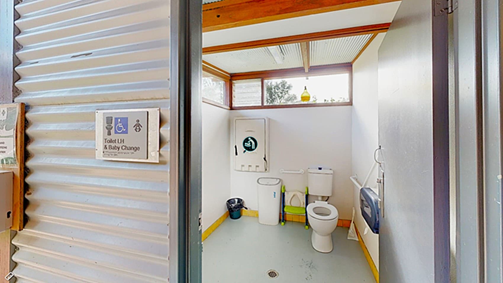 A public restroom with a corrugated metal exterior wall on the left, featuring a sign with symbols. Inside, the restroom has a white toilet on the right, a sink with a mirror above it, and a hand dryer on the wall. A waste bin is on the floor near the sink. The walls are white, and there is a window near the ceiling, allowing natural light to enter. The floor is grey, and the ceiling has wooden beams and white panels.