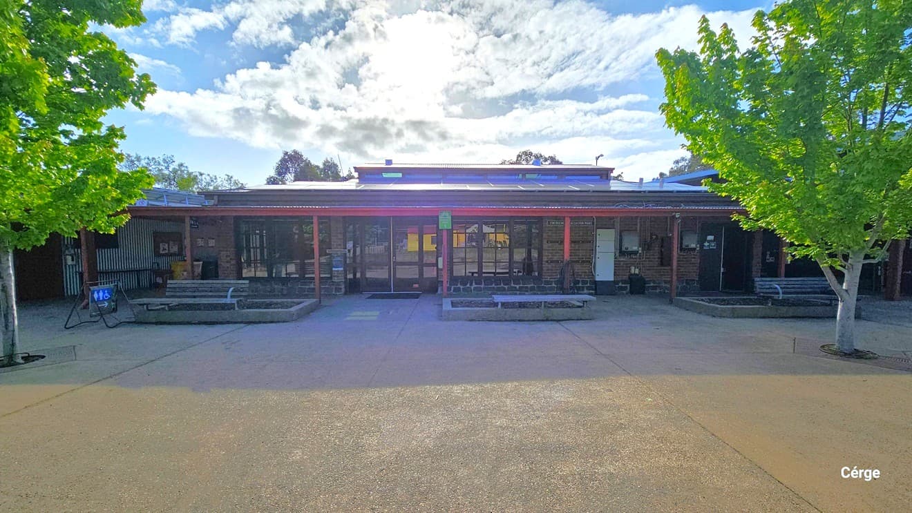 A single-storey building with a flat roof and large windows spans the middle of the image. In front, there are several benches and a paved area. On the left and right, there are green trees with full foliage. The sky is partly cloudy, and the sun is visible above the building, casting shadows on the ground.