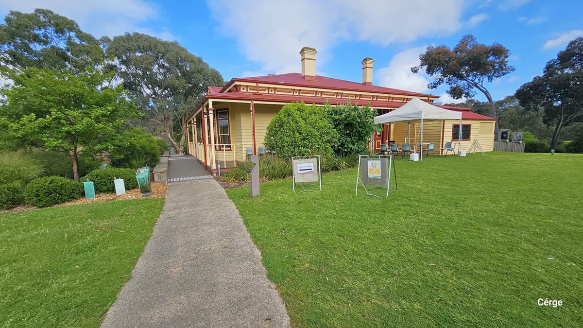 A yellow wooden house with a red roof and two chimneys is situated in a grassy area. On the right, a white canopy is set up with chairs underneath. Two signs are displayed on the grass near the canopy. A paved path leads to the house, bordered by green shrubs and trees. The background features more trees, creating a lush, natural setting.