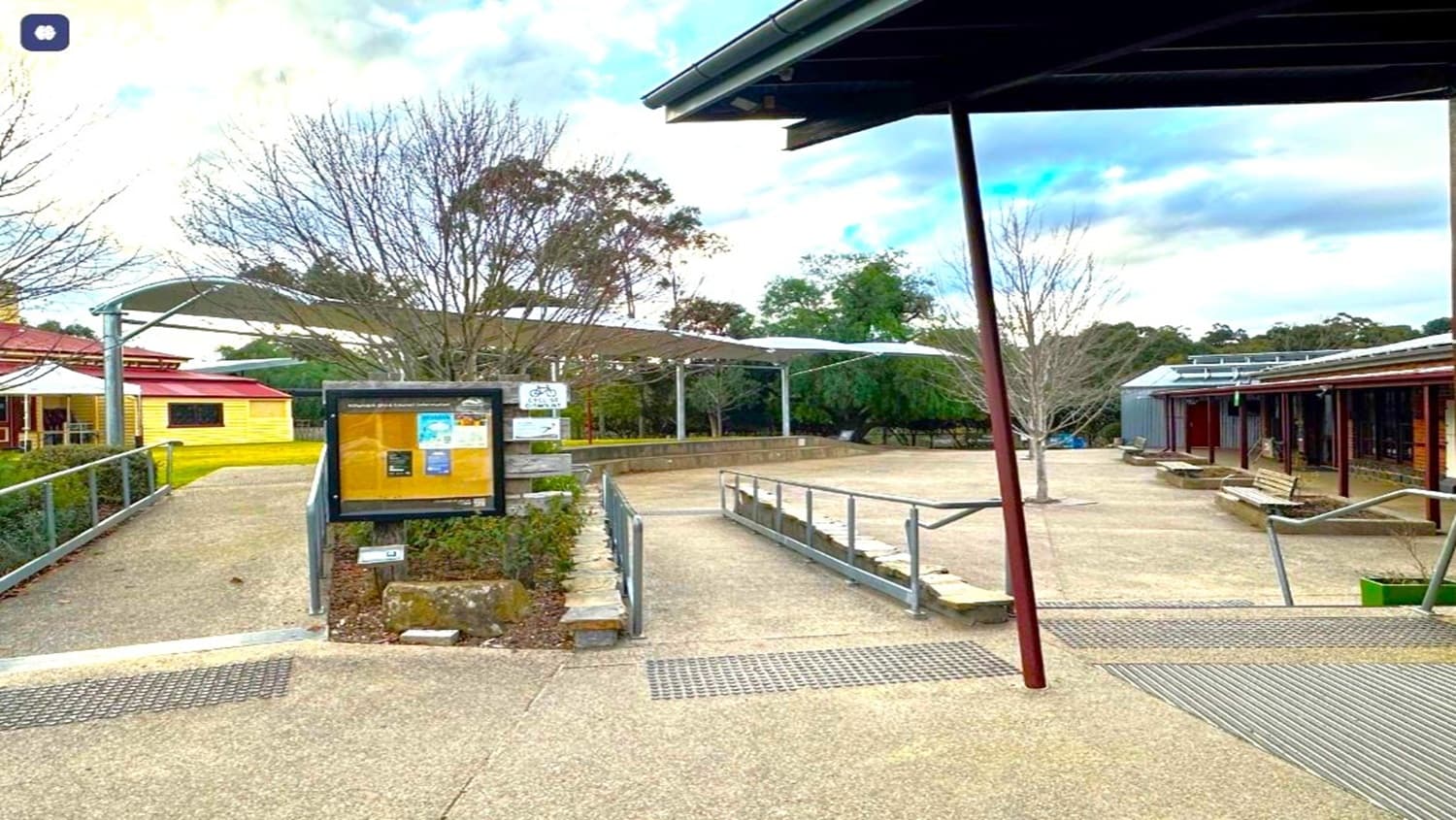 An outdoor area of the community center, featuring accessible ramps, handrails, and a notice board. The space is surrounded by buildings with covered walkways, some trees (mostly bare), and shade structures overhead. The sky is partly cloudy and the setting is calm, with no people visible.