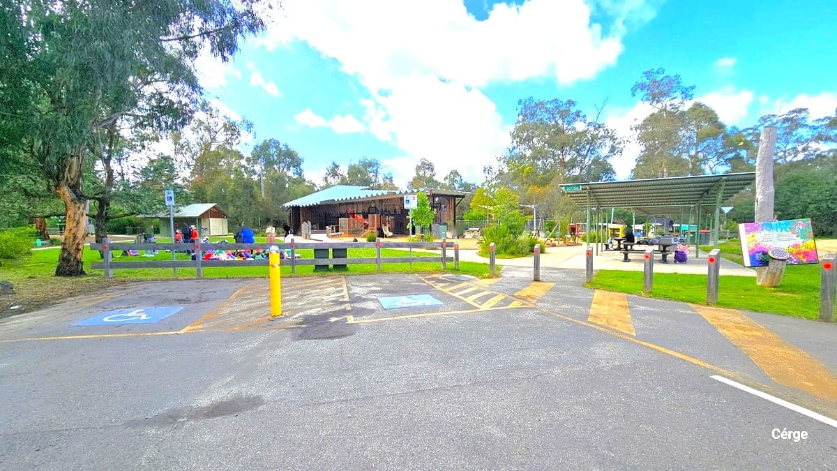 A concrete parking area featuring two accessible car parking spaces with clear indications, accompanied by yellow markings and a yellow bollard. The parking area is bordered by bollards on the right and fenced in the middle. In front lies a generous grassy expanse where individuals are seated or standing beneath the shade of a tall tree. On the right, a shaded region offers picnic tables. Beyond, is the park.